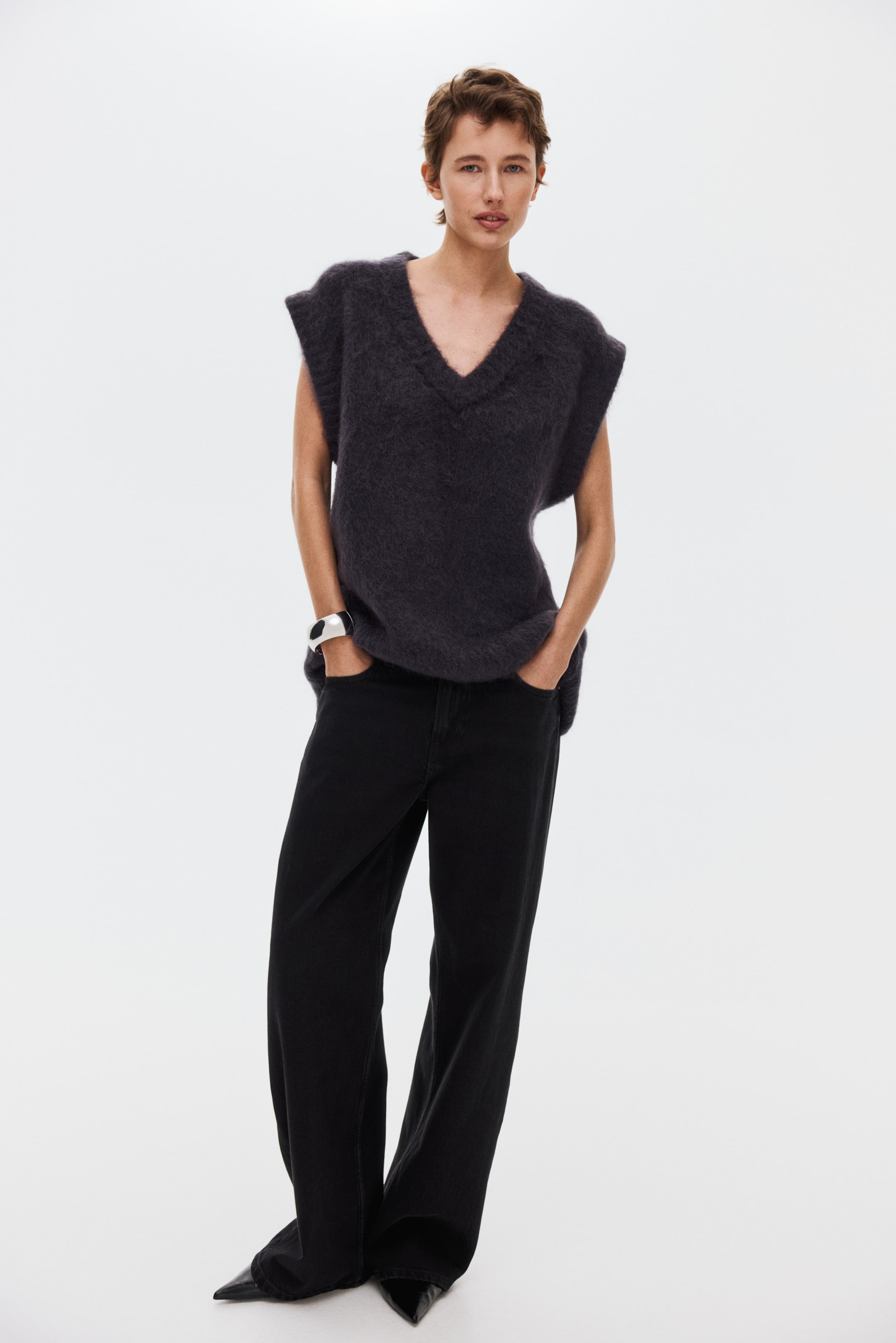 Ampliar la imagen: Se presenta desde un ángulo frontal un chaleco de punto peludo color carbón profundo con escote en V; cuenta con sisas de corte amplio y un ajuste relajado. Cae suelto sobre unos jeans negros de pierna ancha.