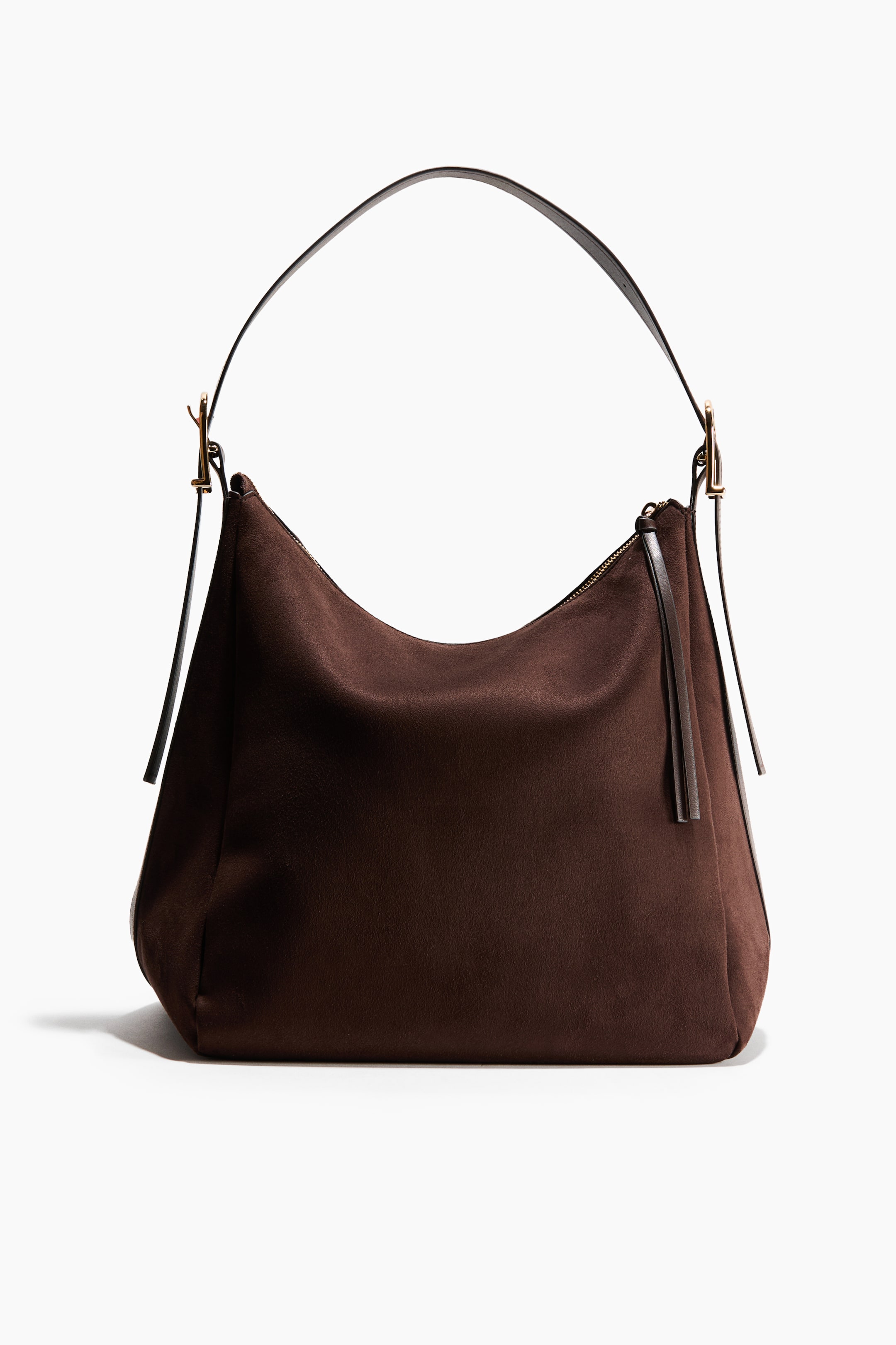 View larger image: A dark brown suede shoulder bag is shown from the front, featuring a slouchy hobo silhouette with a curved top and a single flat leather strap attached with gold-tone metal buckles. The bag features a top zipper closure with a long leather tassel pull and dangling strap ends on the sides.
