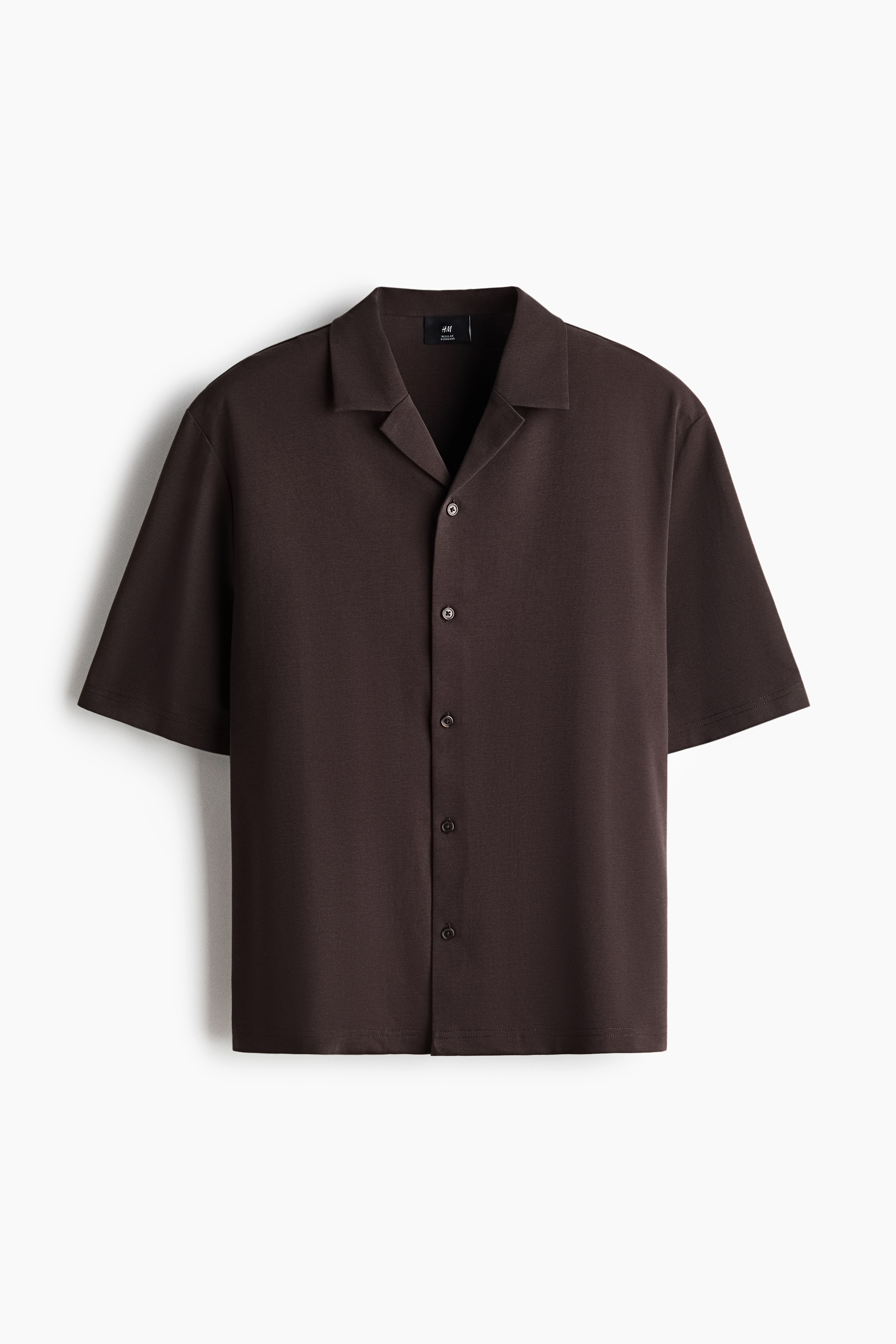 A dark brown, short-sleeve jersey shirt with a resort collar is being worn open by the model. The shirt is layered over a white crew-neck t-shirt and paired with dark trousers.