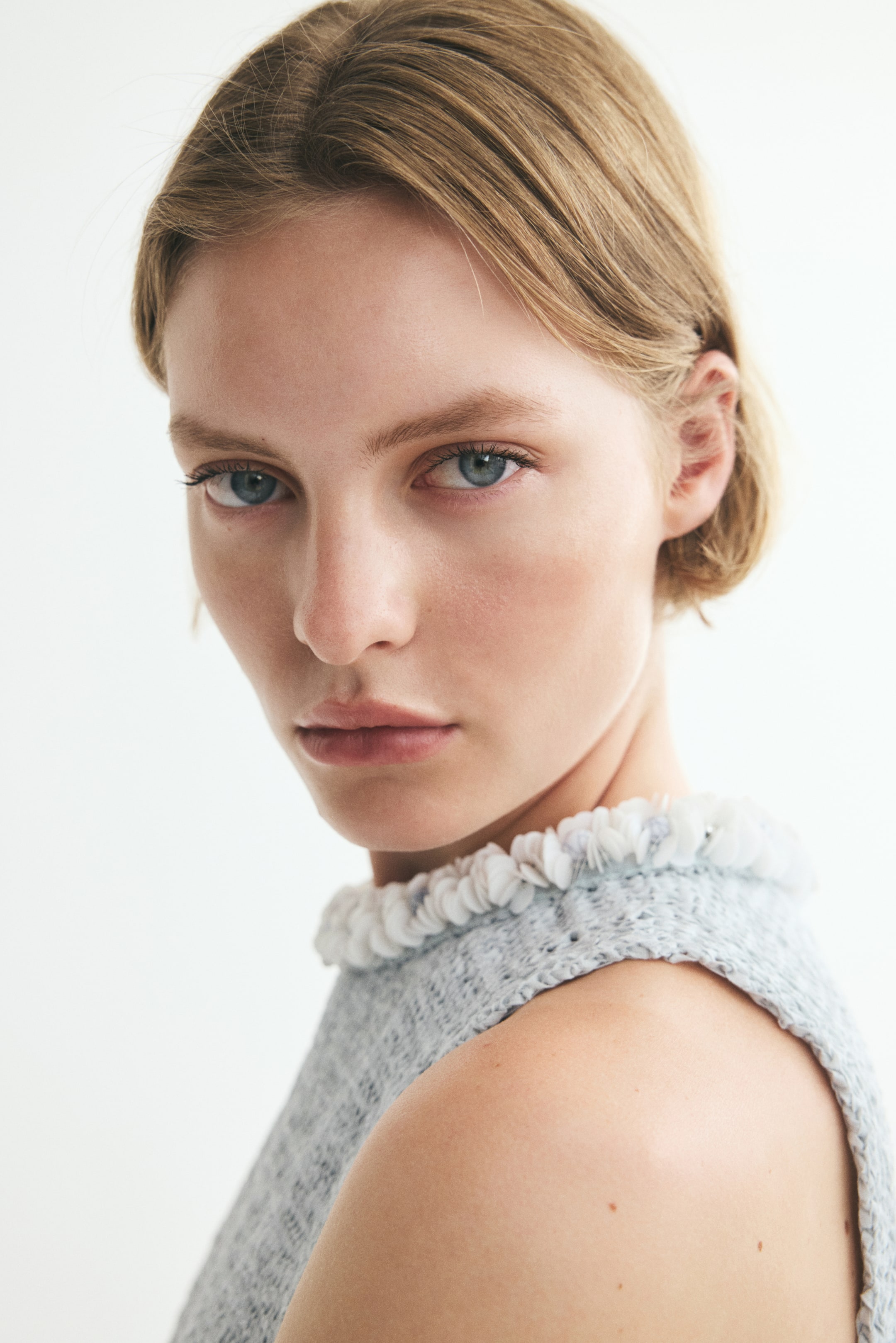 Zobraziť väčší obrázok: Modelka má na sebe bezrukávový top z textúrovaného úpletu s okrúhlym výstrihom zdobeným flitrami a korálkami. Viditeľné je modelkino odhalené rameno, ktoré zvýrazňuje bezrukávový dizajn topu.