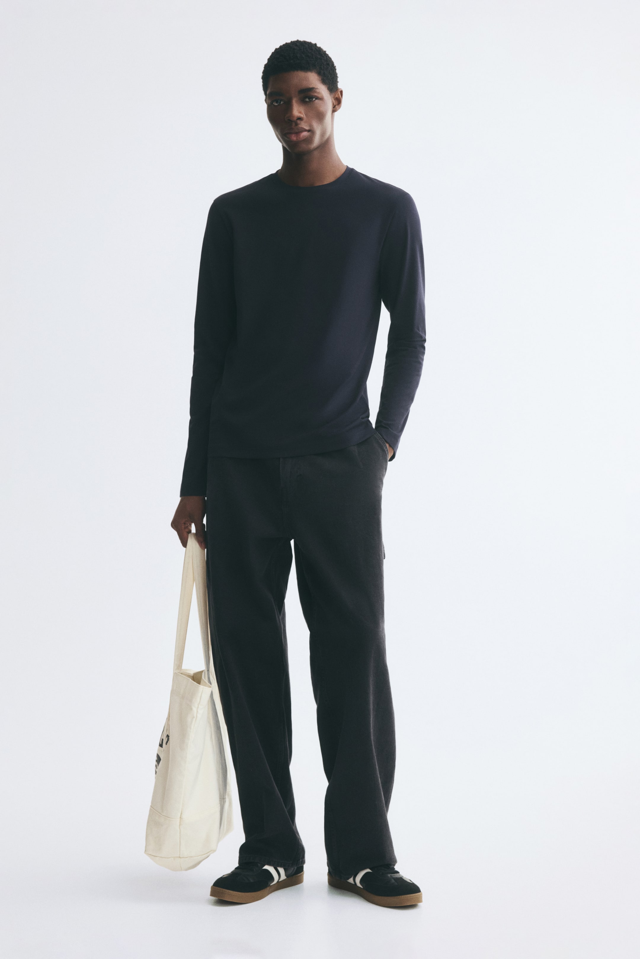 Größeres Bild anzeigen: Das Model trägt ein langärmeliges T-Shirt mit Rundhalsausschnitt in dunklem Marineblau, kombiniert mit einer locker geschnittenen dunklen Hose. Eine cremefarbene Canvas-Tote-Bag wird in einer Hand getragen und rundet das Outfit ab.