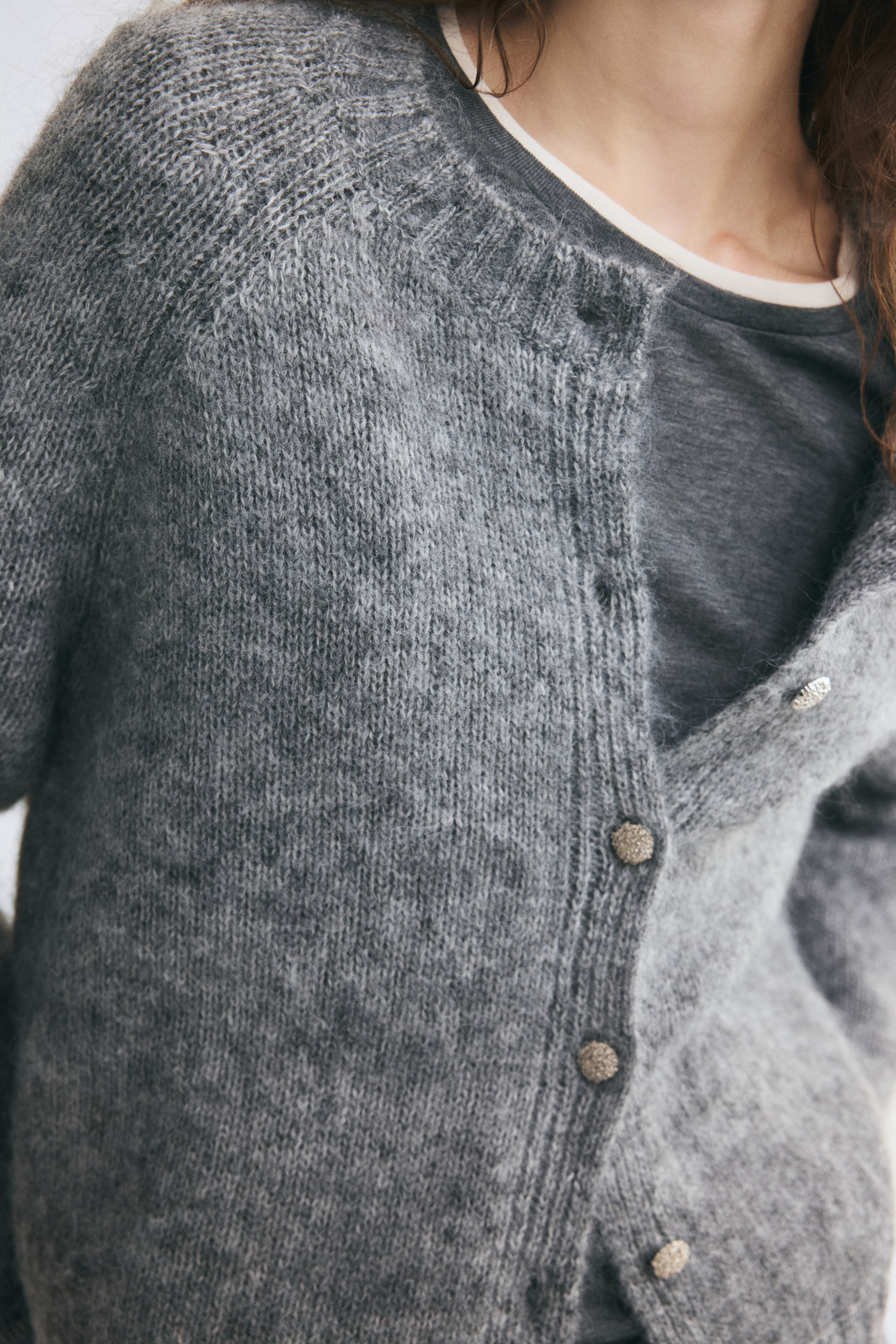 Voir une image plus grande: Un cardigan en tricot doux gris, doté d’un col rond bordé de côtes et de manches raglan, est porté par le mannequin. Le cardigan est stylisé partiellement boutonné avec des boutons métalliques texturés, superposé sur un t-shirt à col rond gris foncé.