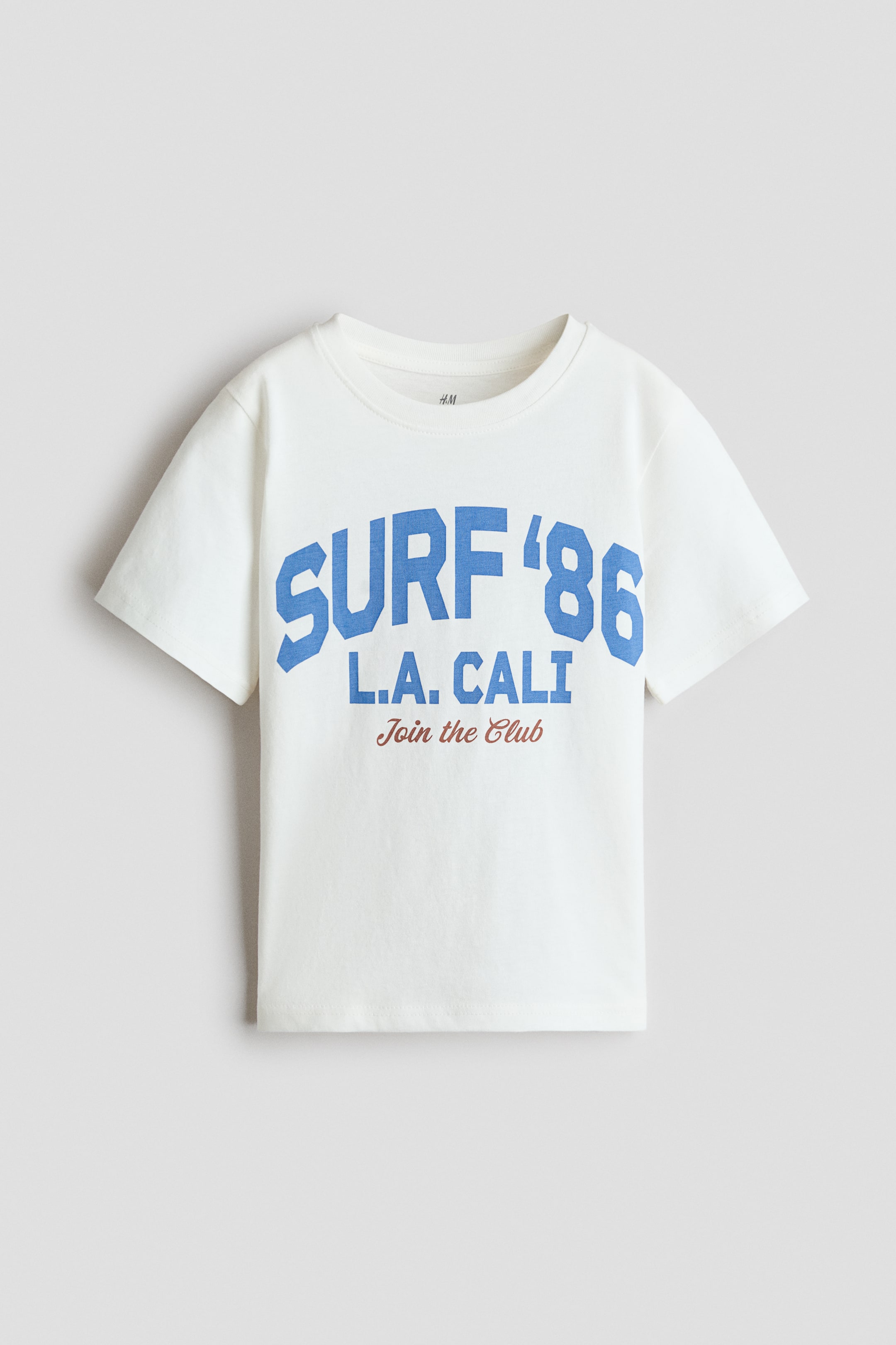 View larger image: An off-white t-shirt in soft cotton jersey is displayed from the front. It features ribbing around the neckline, dropped shoulders, and a straight-cut hem, with an arched "SURF '86" in blue, "L.A. CALI" in blue, and "Join the Club" in brown printed on the front.