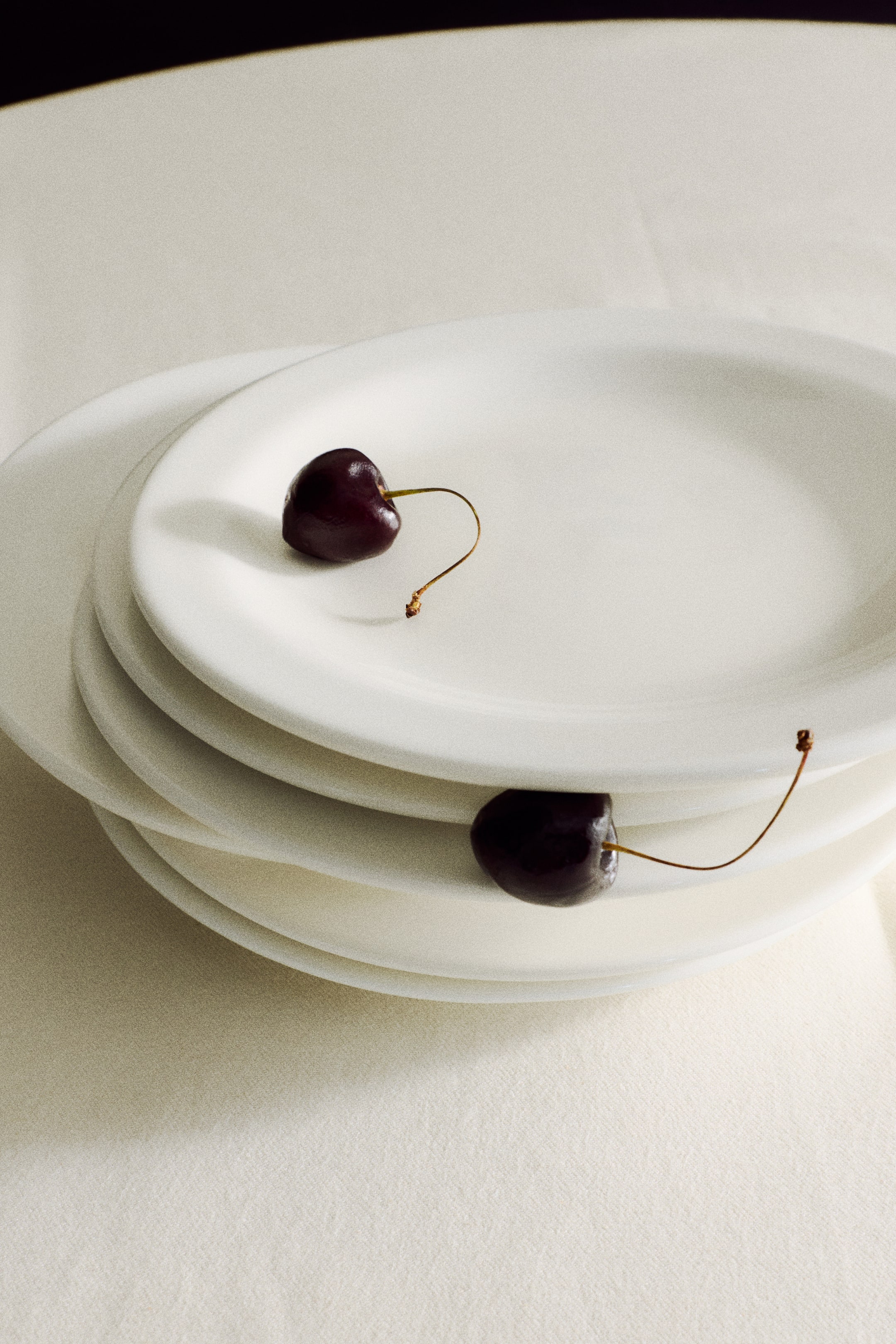 Agrandir l'image: Une pile d’assiettes en céramique blanches et rondes repose en léger décalage, mettant en valeur leur finition mate et lisse ainsi que leurs bords relevés. Une cerise d’un violet foncé avec sa tige est posée sur l’assiette du dessus, tandis qu’une autre est glissée entre les deux premières assiettes, sa tige dépassant vers l’extérieur.