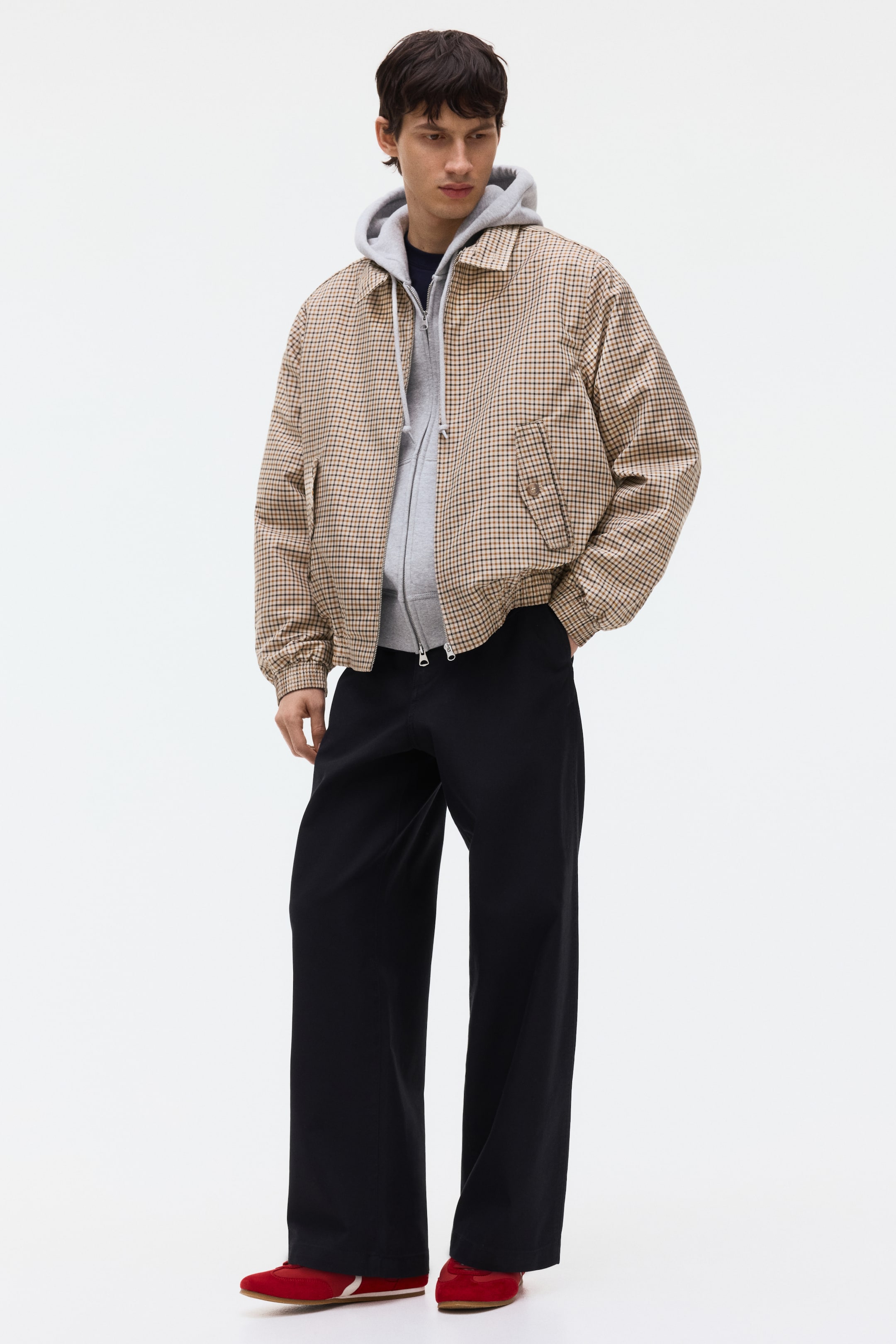 Grösseres Bild ansehen: Ein tan- und dunkel kariertes Jackett wird vom Model getragen und über einem grauen Kapuzenpullover geschichtet. Es präsentiert einen Stehkragen mit Knöpfen, eine teilweise geöffnete Front, diagonale Klappentaschen mit Knöpfen und gesmokte Elastik an den Bündchen und am Saum; das Model steckt eine Hand in eine Jackentasche.