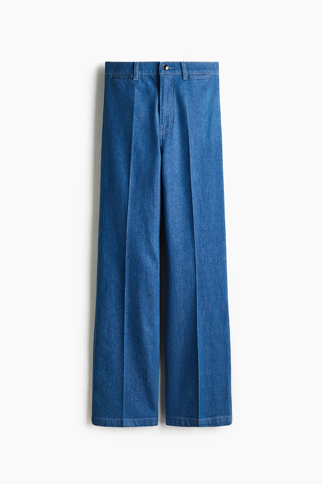 VOORZIJDE: Blauwe denim broek heeft een wijd-uitlopende snit met opvallende plooien aan de voorkant. Ze zijn voorzien van een eenvoudige tailleband met een knoopsluiting en riemlussen. - 1