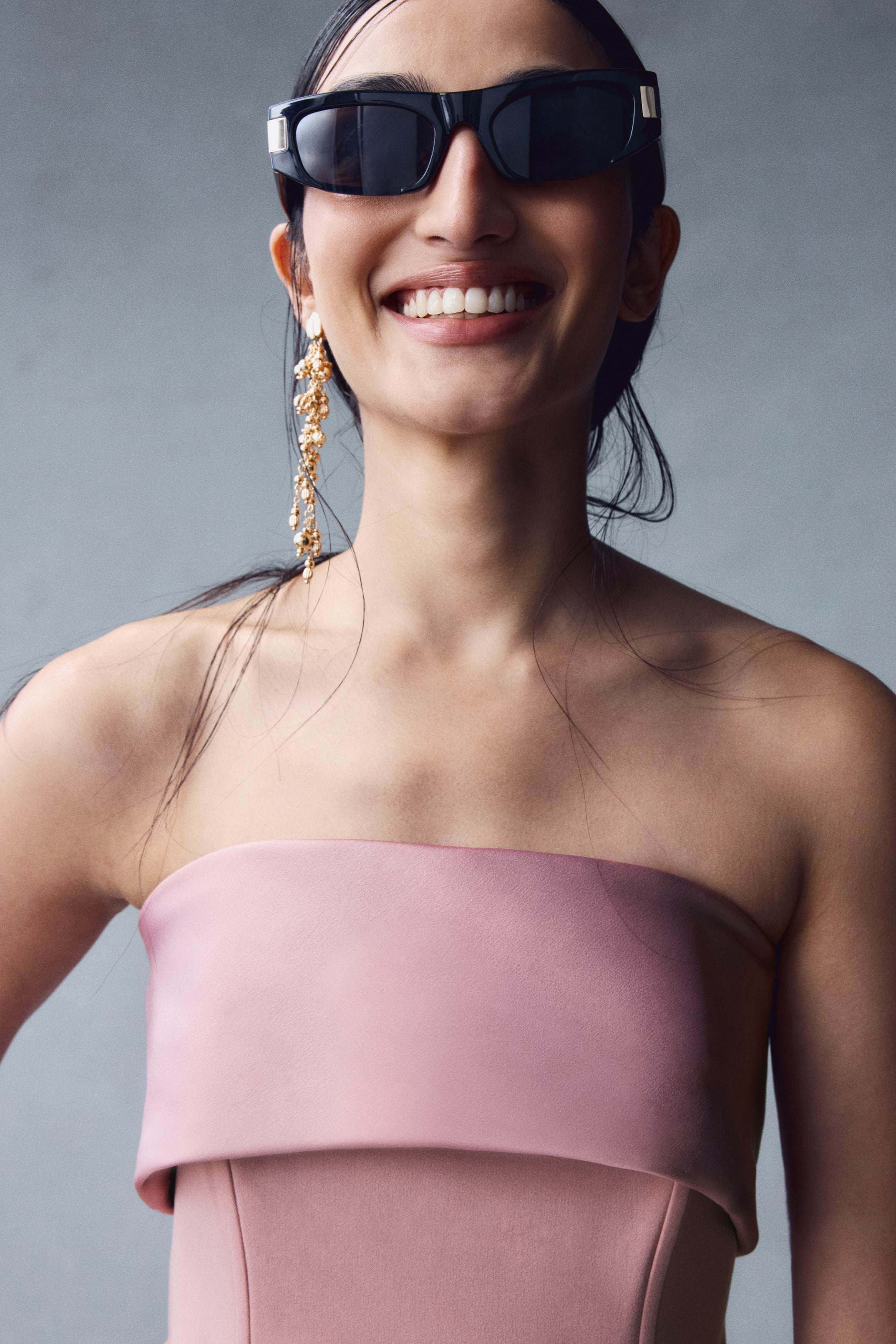 View larger image: A pink bandeau dress featuring a satin foldover top edge and a fitted bodice with vertical structural seams is being worn by the model. The look is styled with black rectangular sunglasses and a long, dangling gold-toned earring.