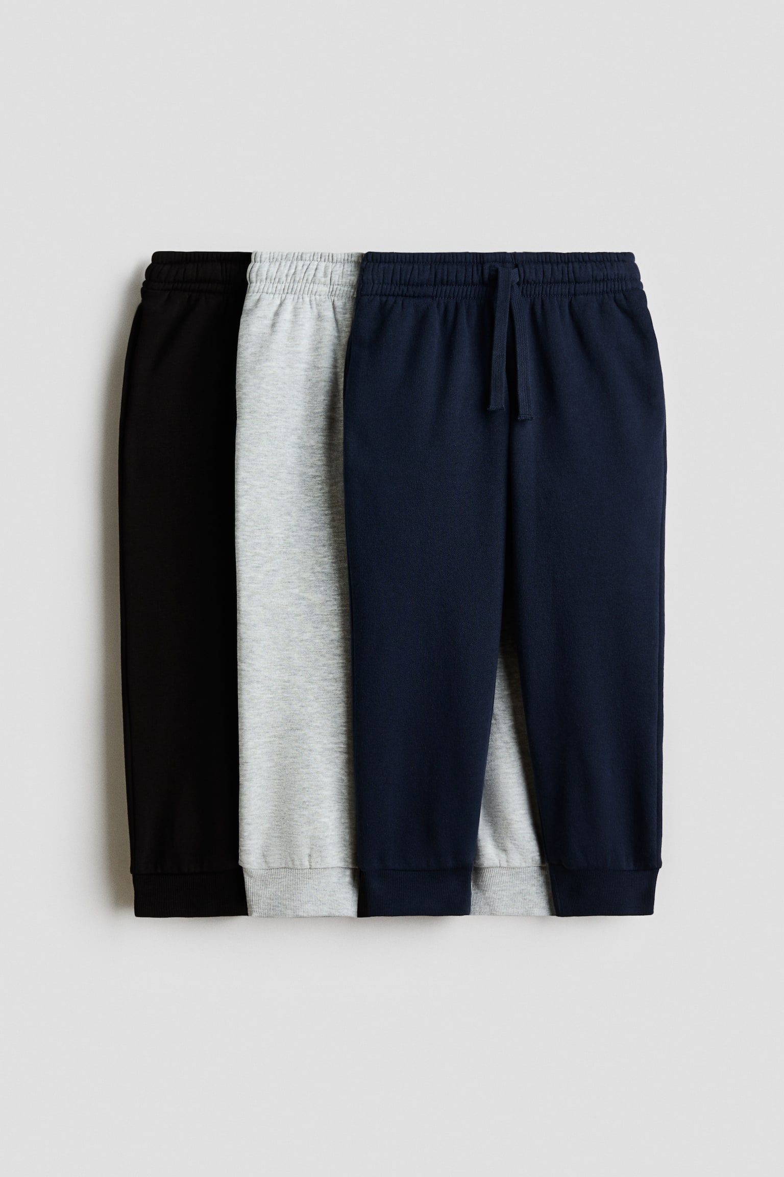 Showing a front view, a three-pack of jogger sweatpants is layered to display black, light grey, and navy blue options. Each pair features an elasticated ribbed waistband and ribbed ankle cuffs, with the foremost navy pair displaying a matching drawstring. - 1