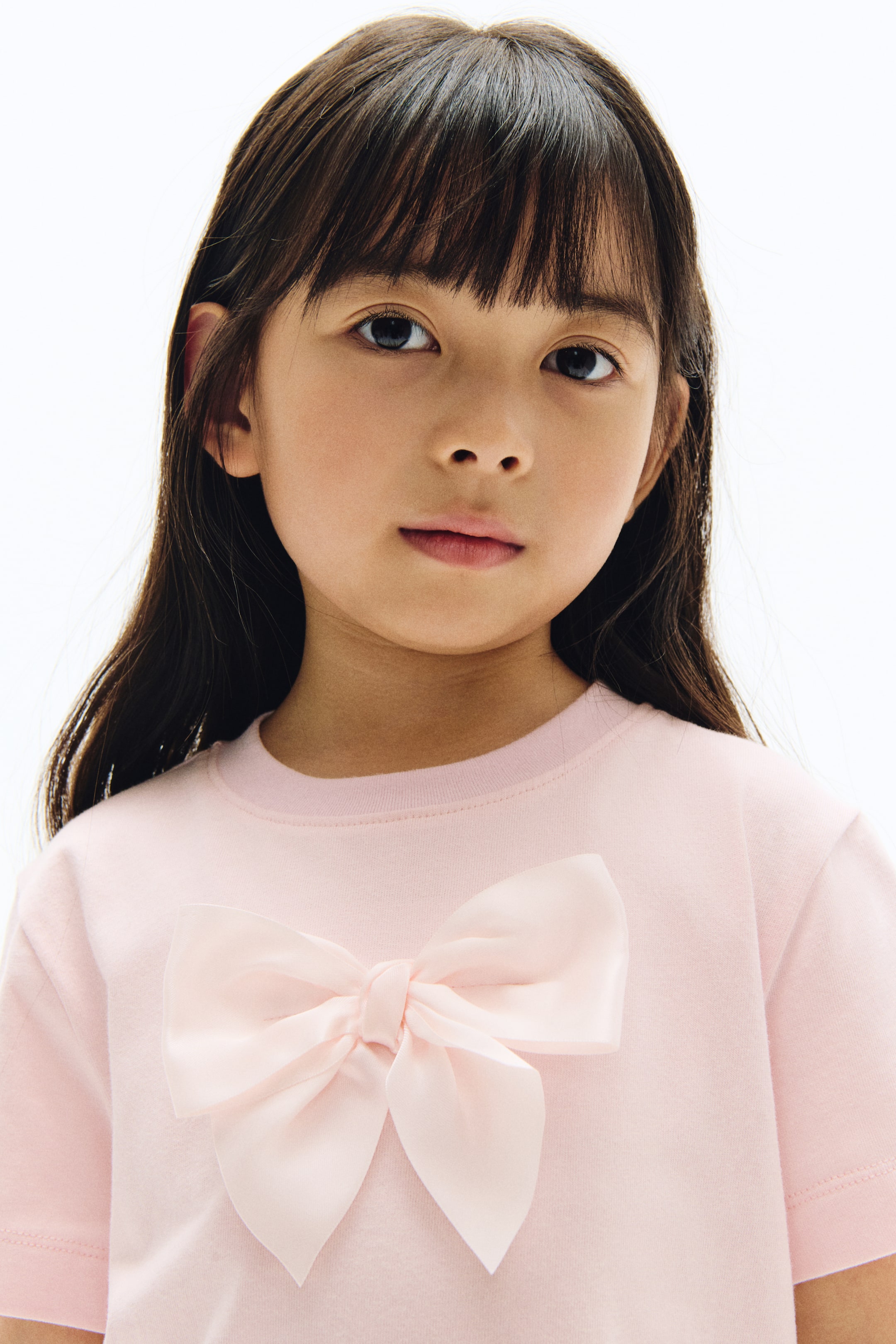 View larger image: A light pink, short-sleeved T-shirt featuring a ribbed crew neck and a large pale pink satin bow attached at the centre of the front.