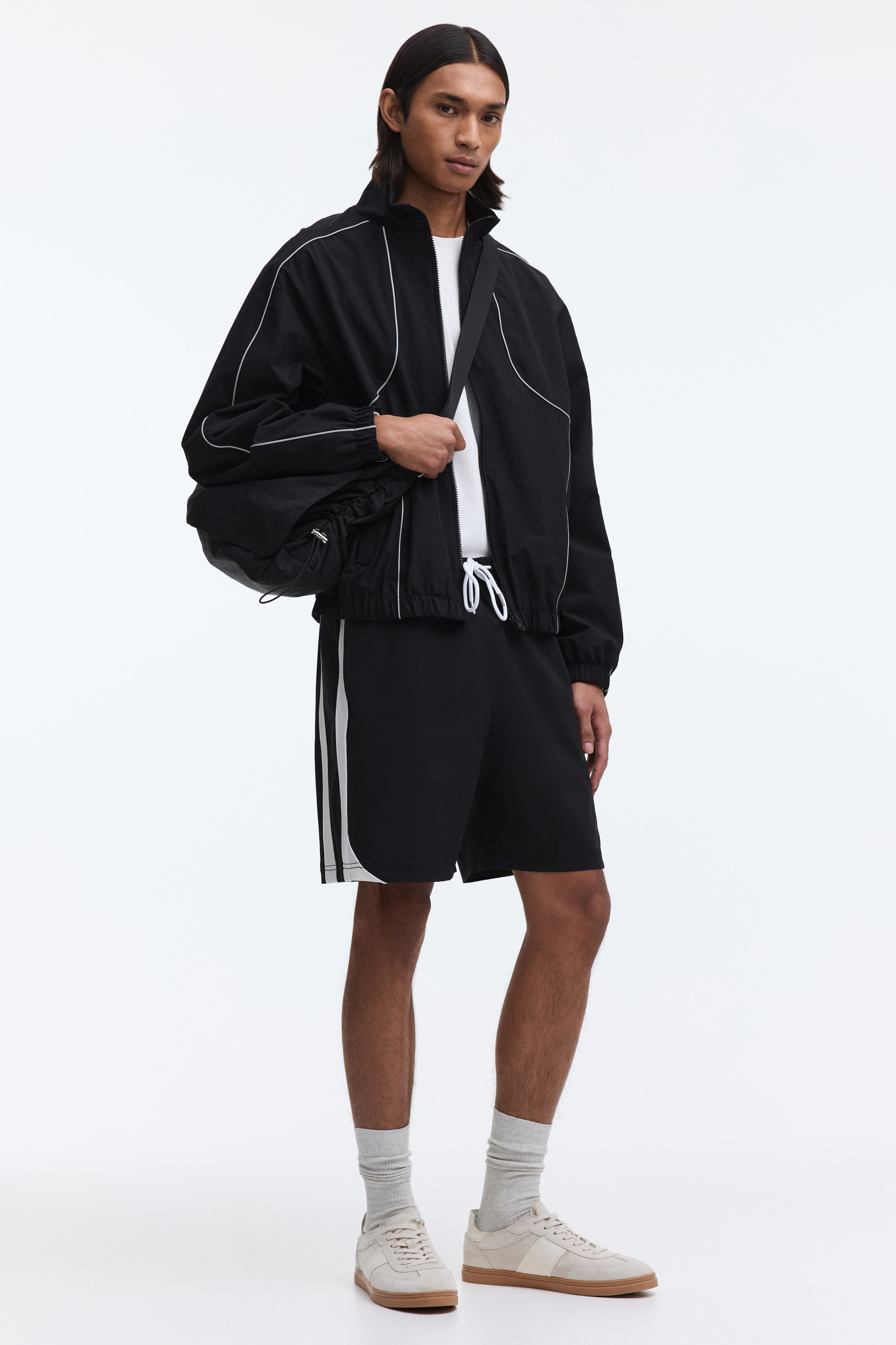 Ampliar la imagen: La modelo lleva puesta una chaqueta deportiva negra con cierre completo al frente, cuello alto, detalles de ribete blanco, y puños y dobladillo elásticos sobre una camiseta blanca. También lleva shorts negros a juego con franjas laterales blancas, y un bolso de hombro negro sobre la chaqueta.