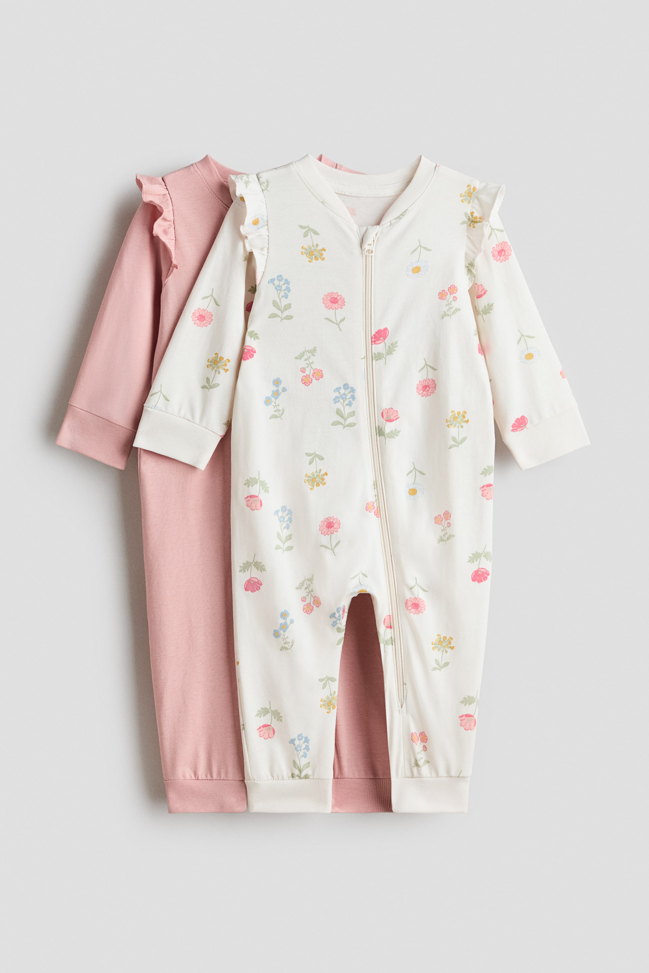 View larger image: Two sleepsuits are presented front-on, with one layered slightly behind the other. The front sleepsuit is off-white with a colourful floral print, featuring long sleeves with ruffles at the shoulders, a full-length front zip and ribbed cuffs and hems. The second sleepsuit, visible behind, is a solid dusty pink and also features long sleeves and ribbed cuffs and hems.