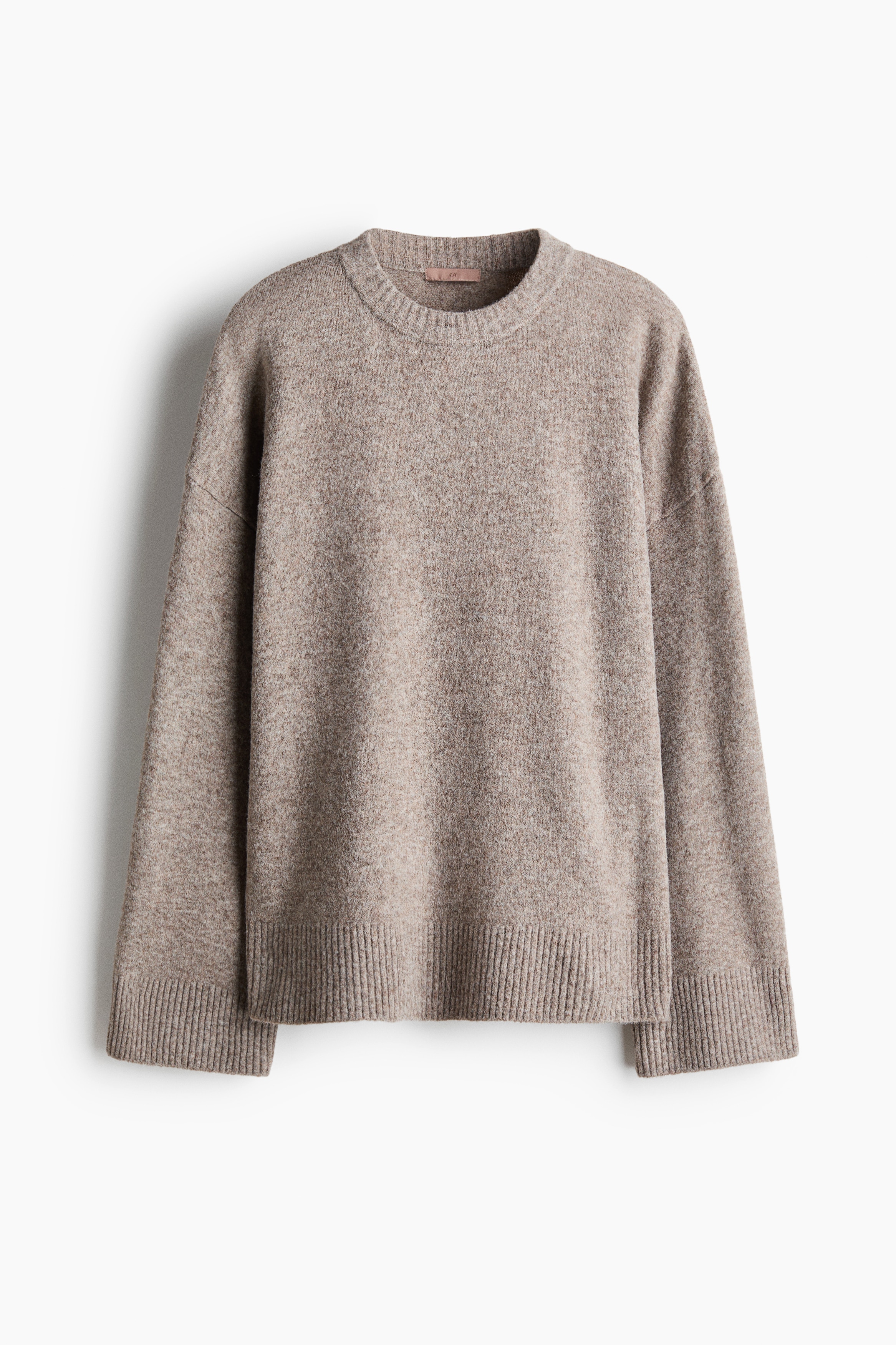 A light-brown knit sweater is shown from the front, featuring a round ribbed neckline, dropped shoulders, and wide ribbed cuffs and hem. The relaxed-fit sweater is styled with a matching knit sweater draped over the shoulders and matching knit trousers.