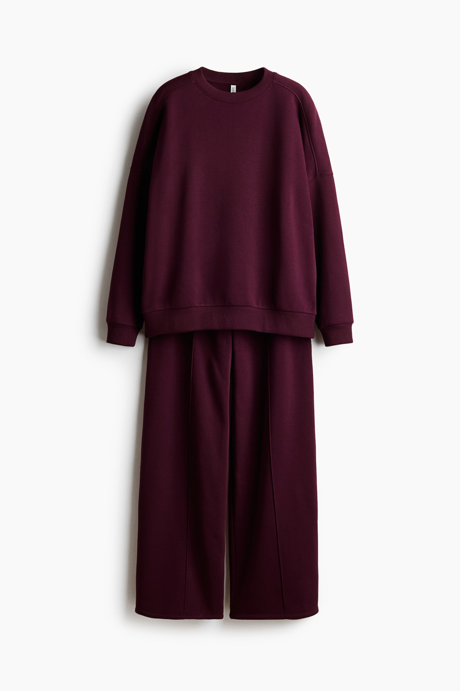 A two-piece burgundy loungewear set is hanging in a front-facing view, featuring a crew-neck sweatshirt and matching wide-leg trousers. The sweatshirt shows dropped shoulders, ribbed cuffs, and a ribbed hem, while the trousers display central vertical seams down the front of each leg.