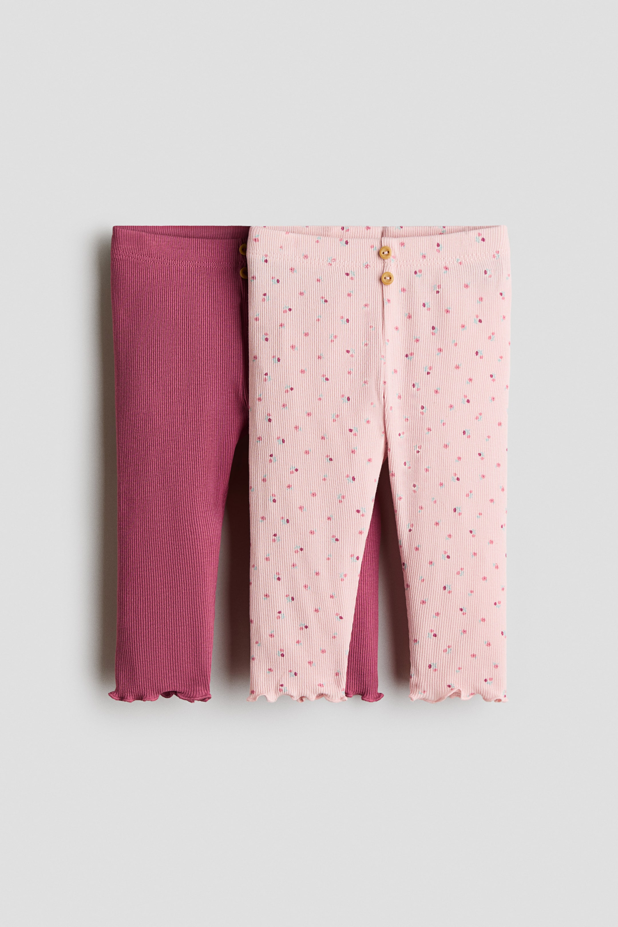 Größeres Bild ansehen: Zwei Paar gerippte Baby-Leggings sind frontal ausgestellt. Das linke Paar ist einfarbig dunkelrosa und hat einen gerüschten Saum, während das rechte hellrosa Paar ein verstreutes Blumenmuster, zwei Holzknöpfe am Bund und einen gerüschten Saum aufweist.
