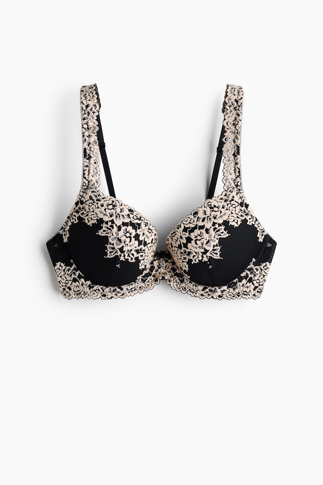 Ein BH präsentiert schwarze, gepolsterte Cups mit einer beigen Blumen-Spitzen-Überlagerung und einem gewellten Rand. Er verfügt über verstellbare schwarze Träger, die ebenfalls mit der passenden beigen Spitze verziert sind, alles aus einer frontalen Perspektive betrachtet. - 1