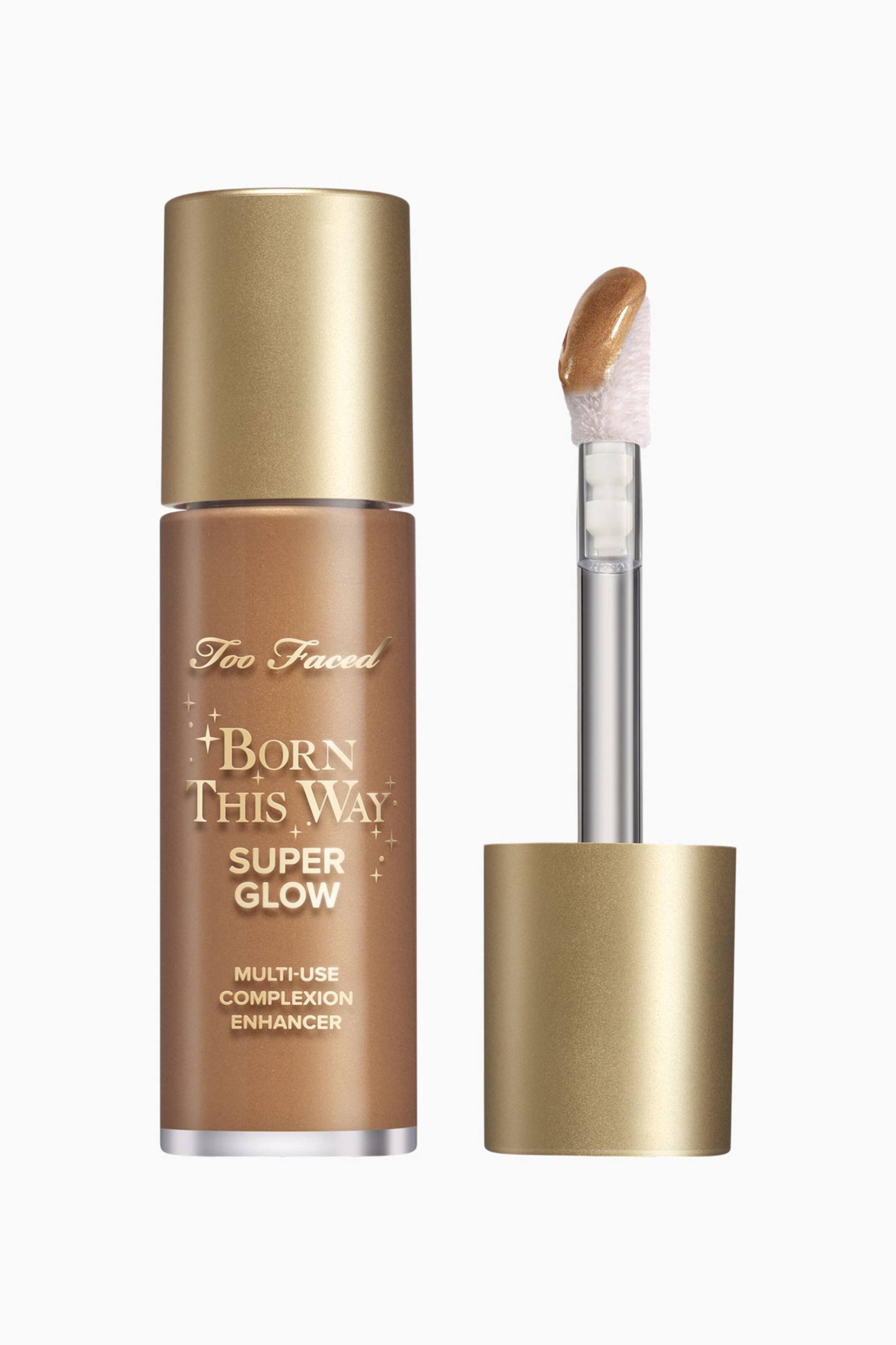 Visa större bild: Flaskan med Too Faced Born This Way Super Glow Multi-Use Complexion Enhancer står upprätt, visar sin bronsfärgade vätska, guldfärgade kork, präglad text och silverband vid basen. Bredvid står den borttagna applikatorn och visar en skimrande gyllenbronsfärgad produkt på sin mjuka spets.