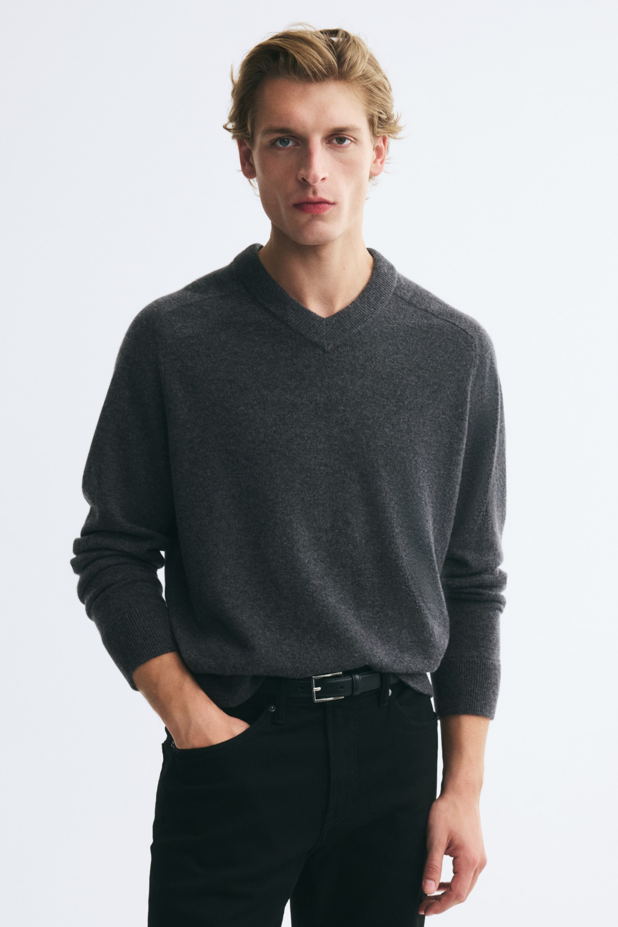 View larger image: A dark grey knit V-neck sweater is worn by the model, who is standing with one hand tucked into the pocket of black jeans. The sweater features a ribbed collar and cuffs and is styled tucked into a black leather belt.