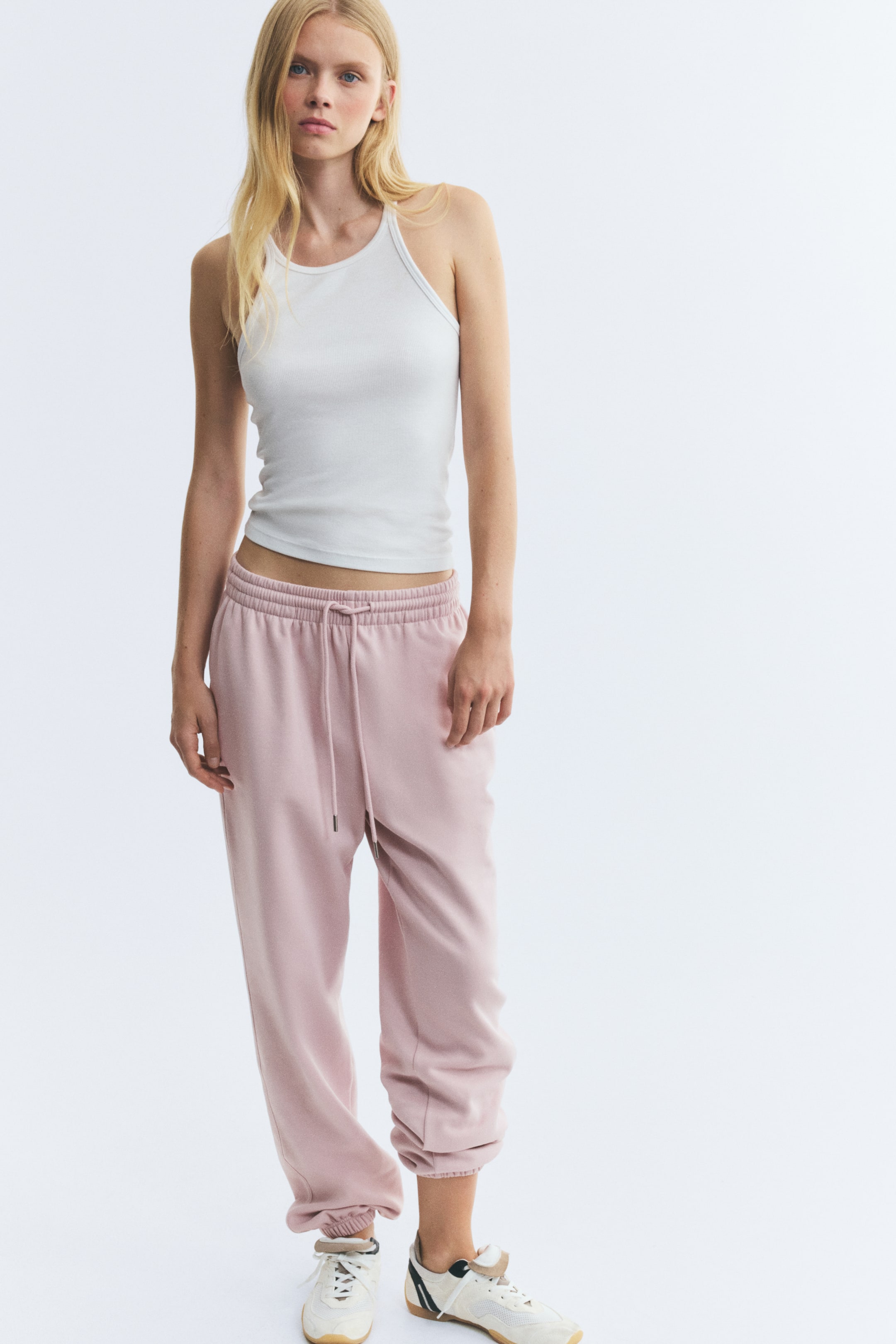 Ver imagen más grande: La modelo lleva un pantalón de chándal en tejido sudadera de color rosa claro, con cintura alta, cintura elástica cubierta y cordón ajustable. El pantalón de corte relajado presenta bajos elásticos en los tobillos y se combina con un top halter blanco y zapatillas blancas rotas.