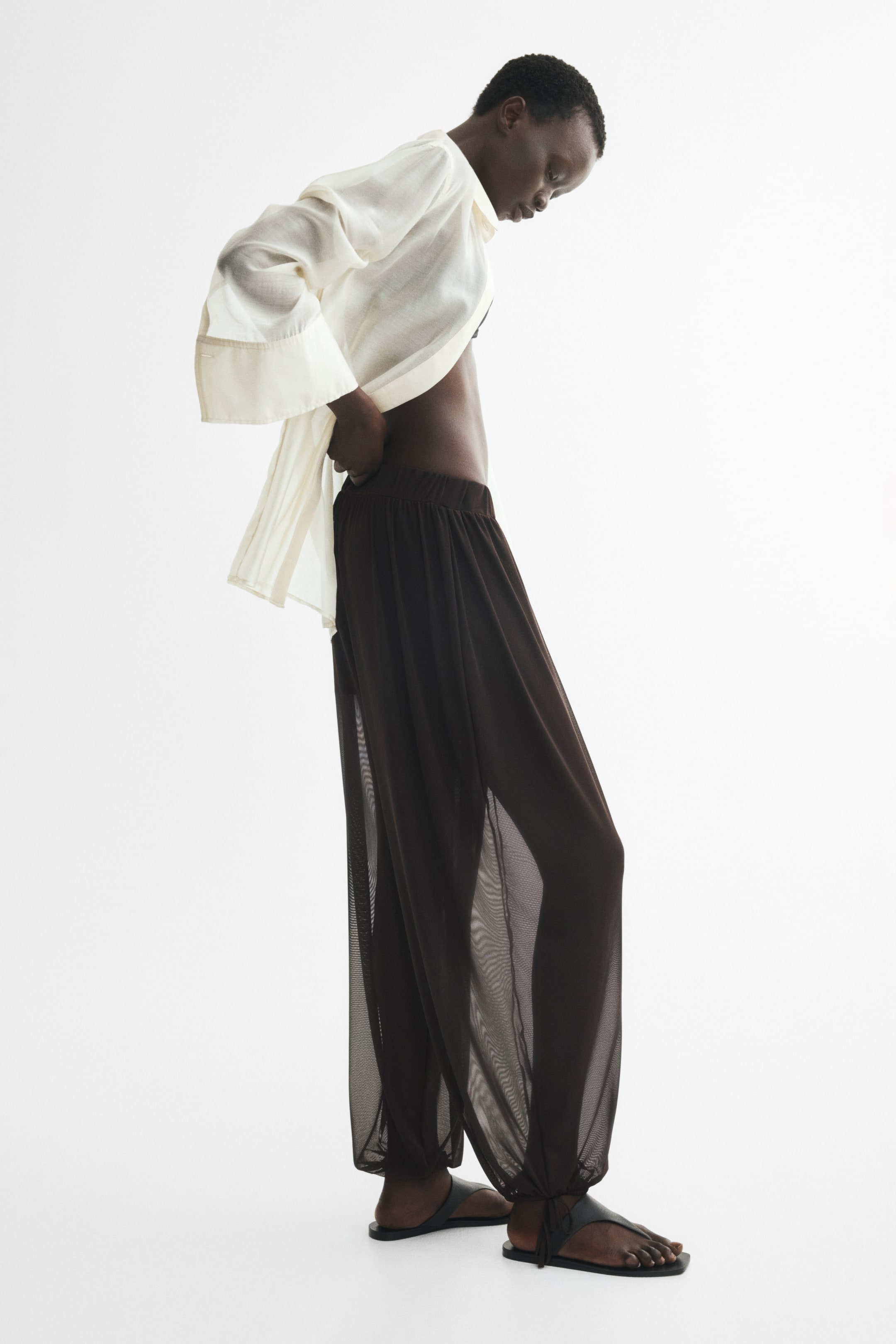 View larger image: Dark brown mesh trousers, designed with a high, elasticated waist, balloon legs featuring covered elastication and drawstrings at the hems, and a partially lined construction creating a sheer effect on the legs, are worn by the model, who is adjusting the waistband.