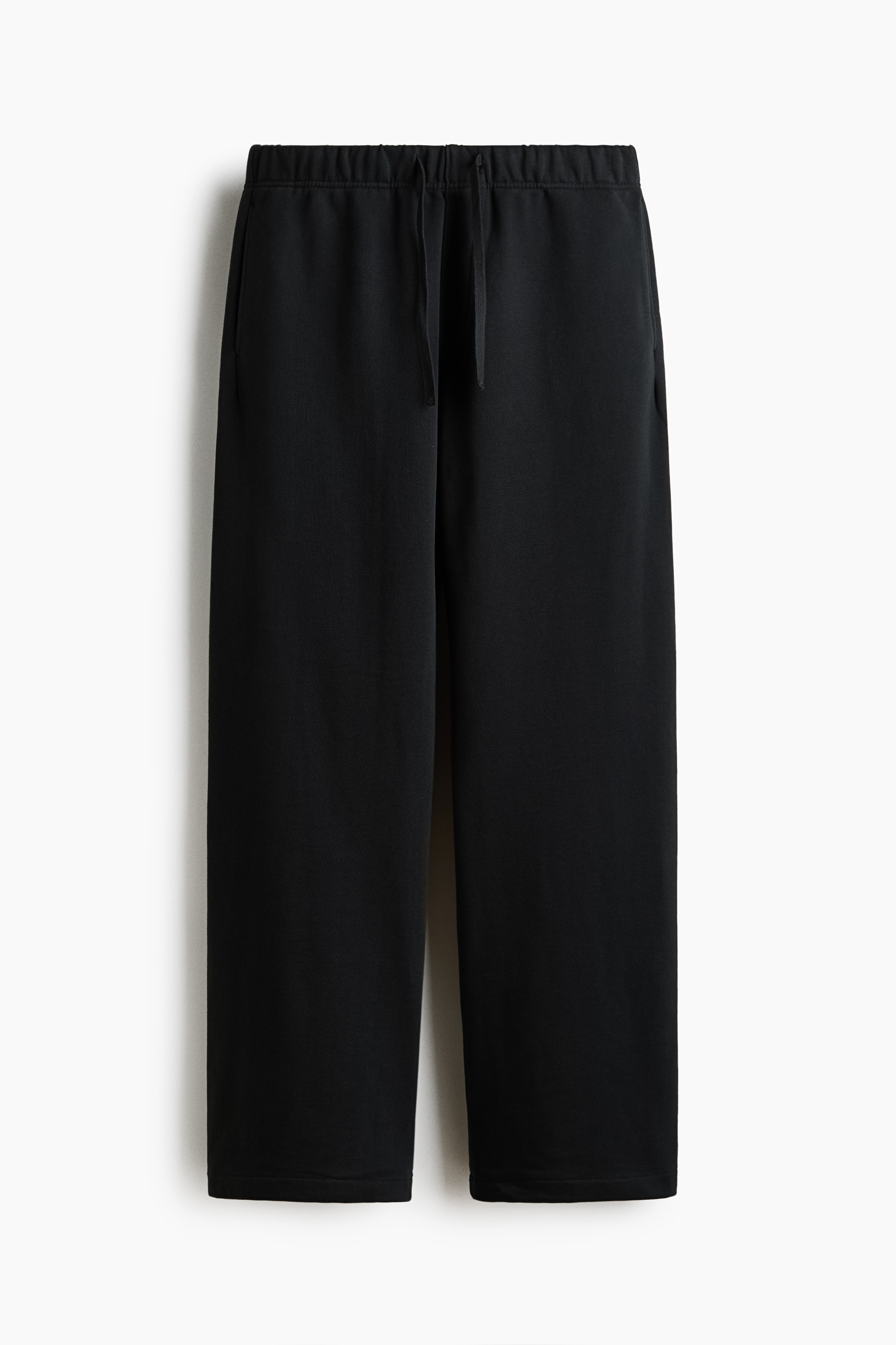 Black track pants with a relaxed fit and straight legs are being worn by the model. The pants, which feature an elasticized drawstring waist, are styled with a dark purple graphic hoodie and black runners.