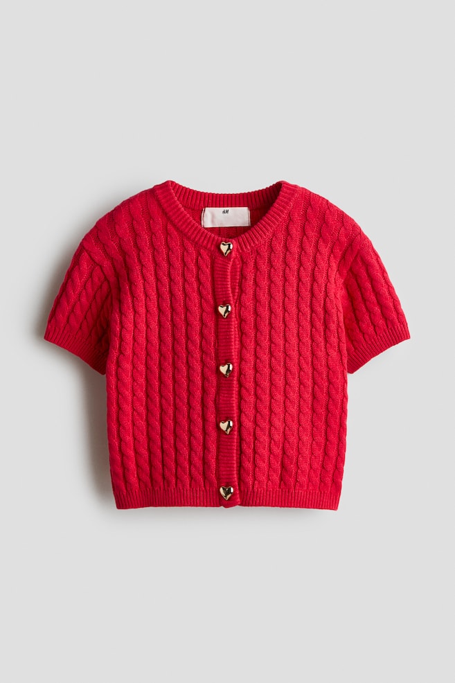A red, short-sleeved cable-knit cardigan is shown front-facing, featuring a ribbed crew neck and five gold-toned, heart-shaped buttons down the placket. The cardigan also features ribbed finishes at the sleeve cuffs and hem. - 1