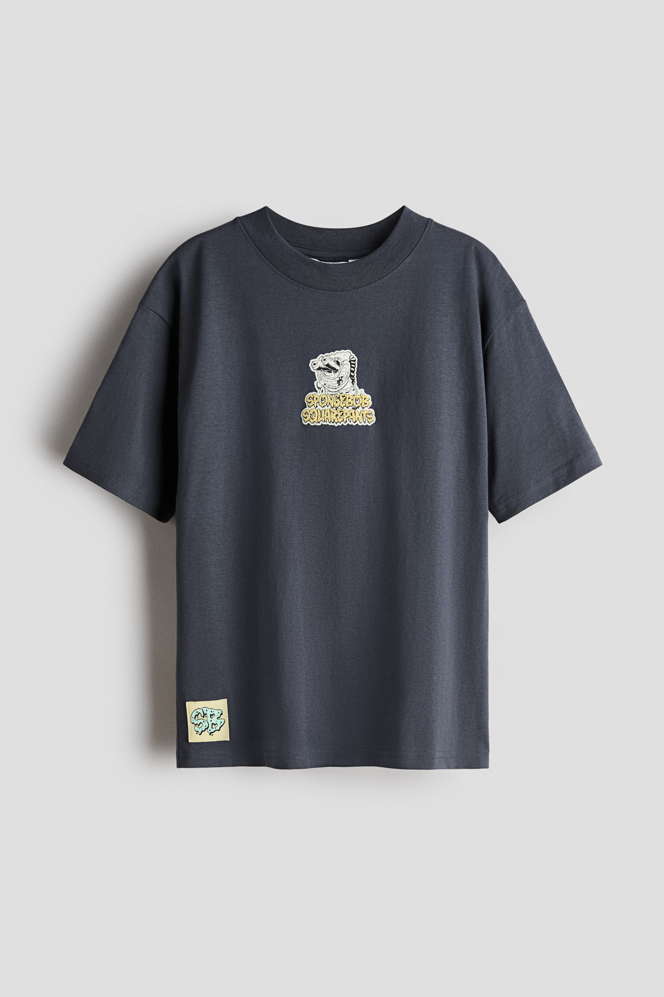 View larger image: The front view showcases a dark grey cotton jersey t-shirt, highlighted by a ribbed trim around the neckline. It offers a relaxed fit with dropped shoulders, adorned with a graphic print on the chest. The print features a cartoon character above the text 'SPONGEBOB SQUAREPANTS'. A small, square yellow patch with a light blue graphic can be seen on the lower left hem.