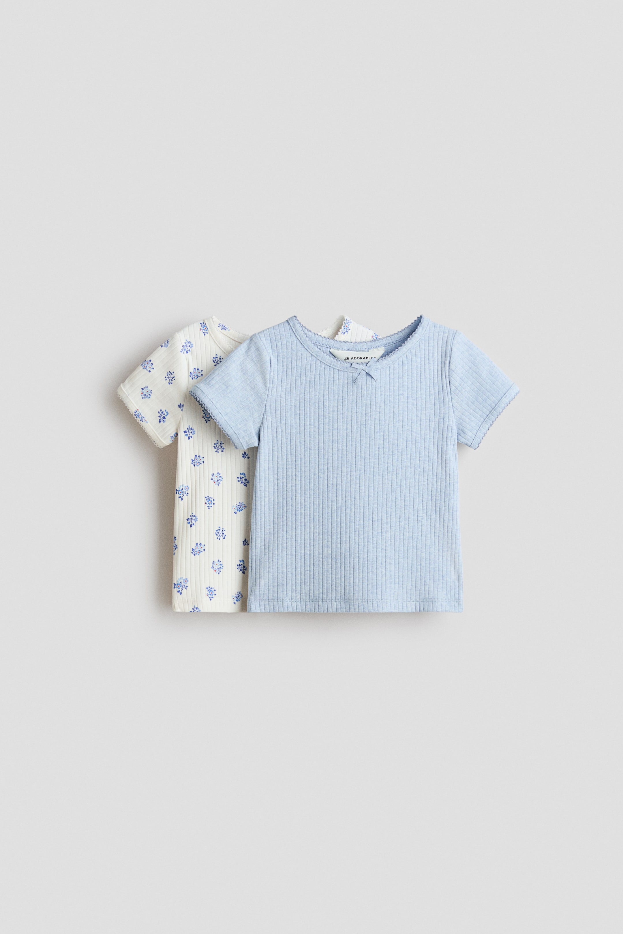 Grotere afbeelding bekijken: Twee H&M Adorables T-shirts met korte mouwen zijn frontaal in beeld. Beide T-shirts zijn gemaakt van geribde jersey, hebben een ronde hals met picotafwerking en een klein satijnen strikje. Eén T-shirt is lichtblauw, het andere is offwhite met een blauw bloemenmotief.