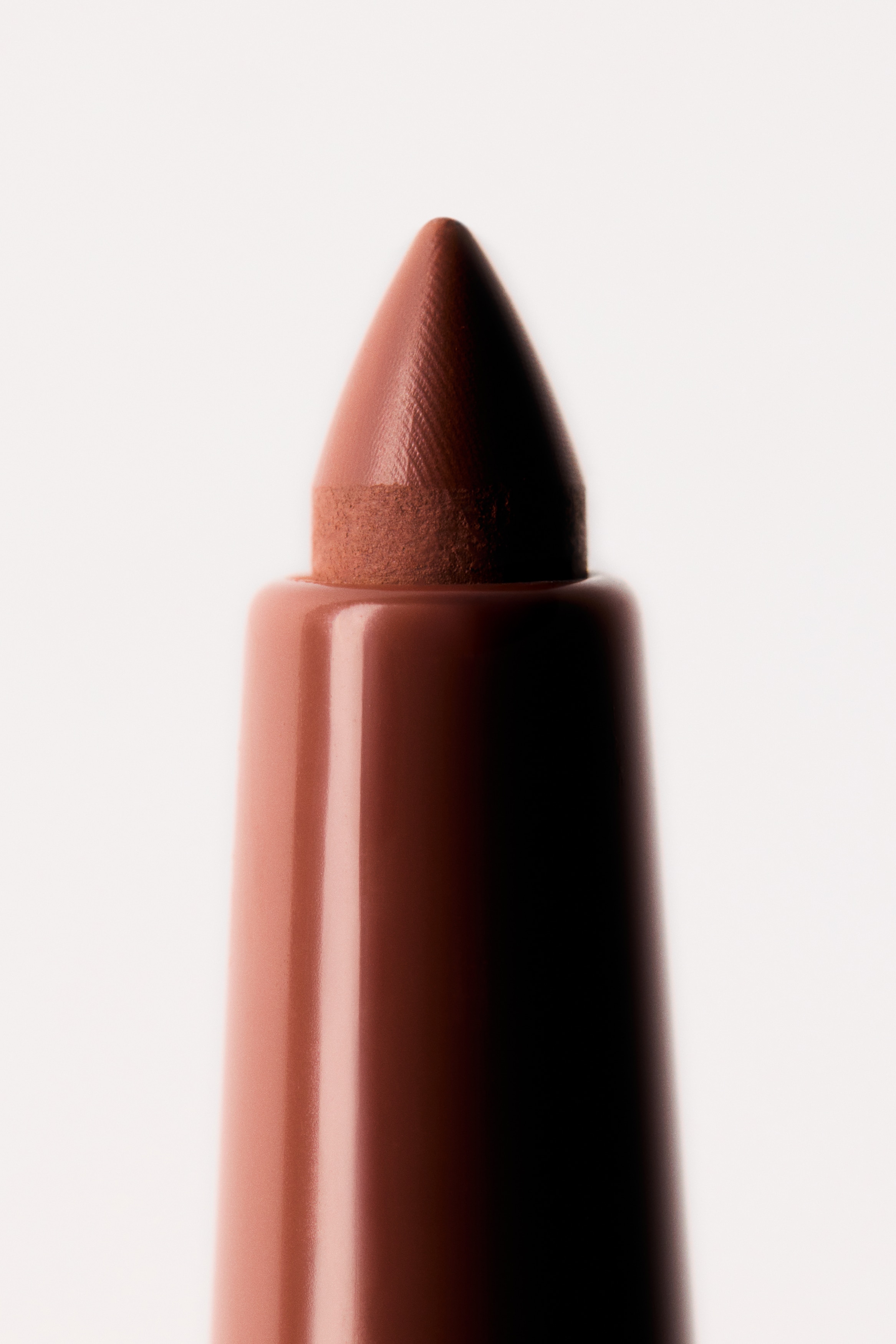 Dva valcovité ceruzky na pery stoja vzpriamene, smerujú dopredu, obe majú lesklý kovový strieborný valec s nápisom "PLUMPING LIP LINER" a "SANDY BLOOM" a hnedým indikátorom farby na spodnej časti. Jedna ceruzka je uzavretá kovovým strieborným viečkom, druhá je otvorená, odhaľujúc špičatý hnedý hrot ceruzky na pery.