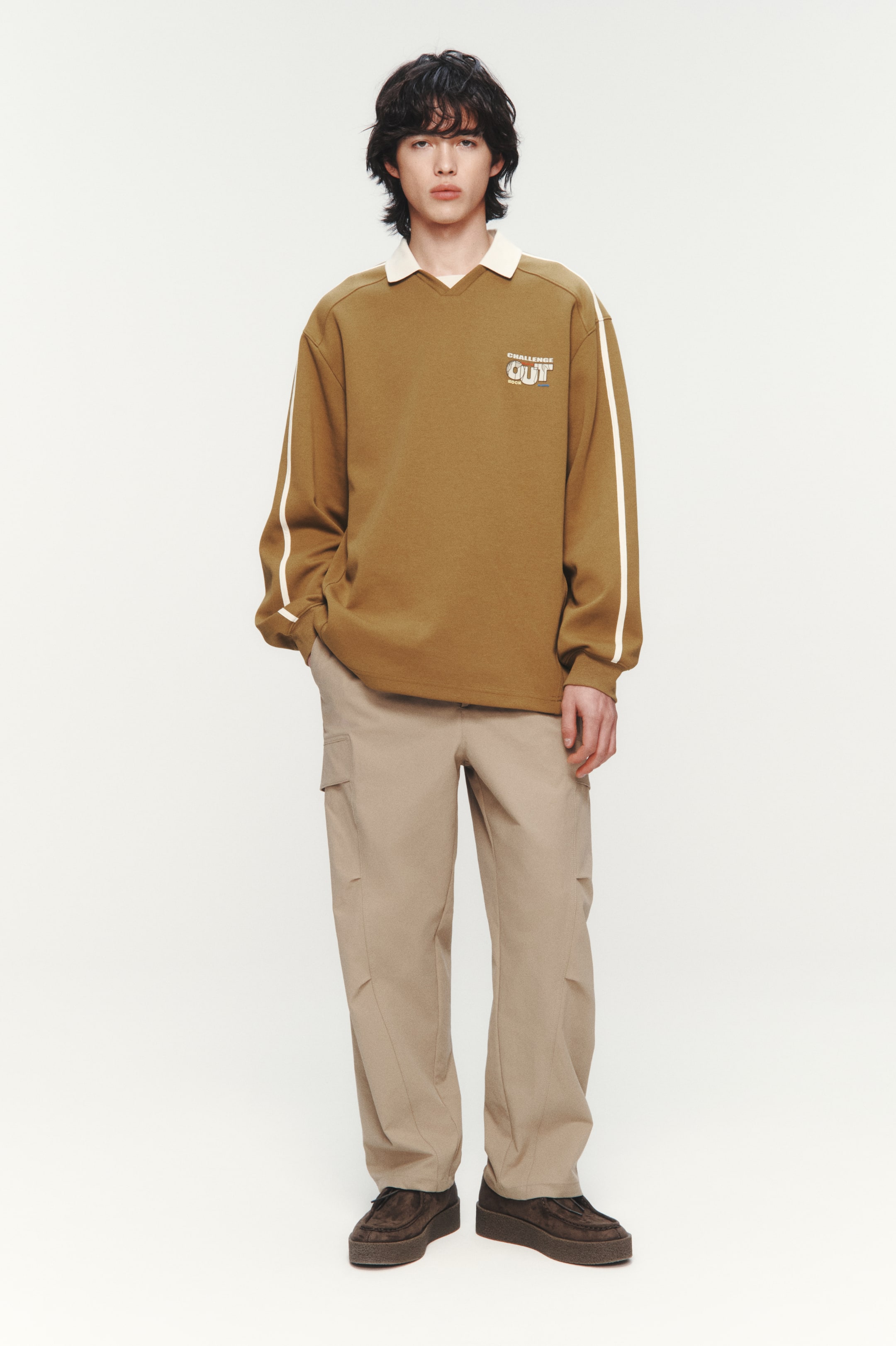이미지 확대 보기: 갈색 긴팔 폴로 셔츠가 소개됩니다. 이 셔츠는 대조적인 흰색 뾰족한 칼라, V넥 오프닝, 그리고 양쪽 소매 바깥쪽에 흰색 줄무늬가 특징입니다. 왼쪽 가슴에는 "CHALLENGE OUT" 텍스트와 그래픽 로고가 나타납니다.