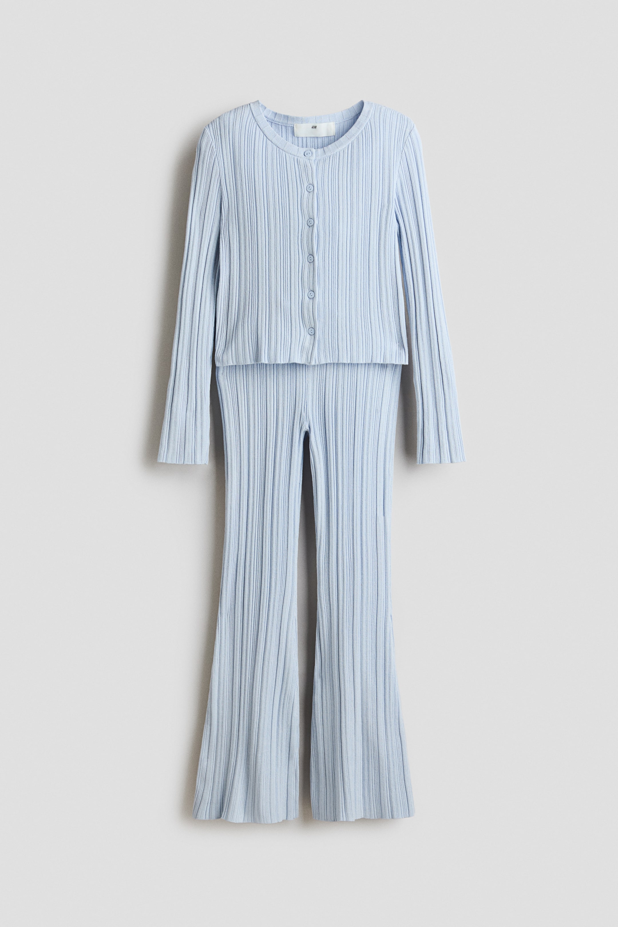 Zobrazit větší obrázek: Světle modrý žebrovaný pletený komplet je zobrazen zepředu; obsahuje kardigan s dlouhým rukávem a kulatým výstřihem se zapínáním na knoflíky vpředu a ladící rozšířené kalhoty. Obě části mají výraznou vertikální žebrovanou strukturu.