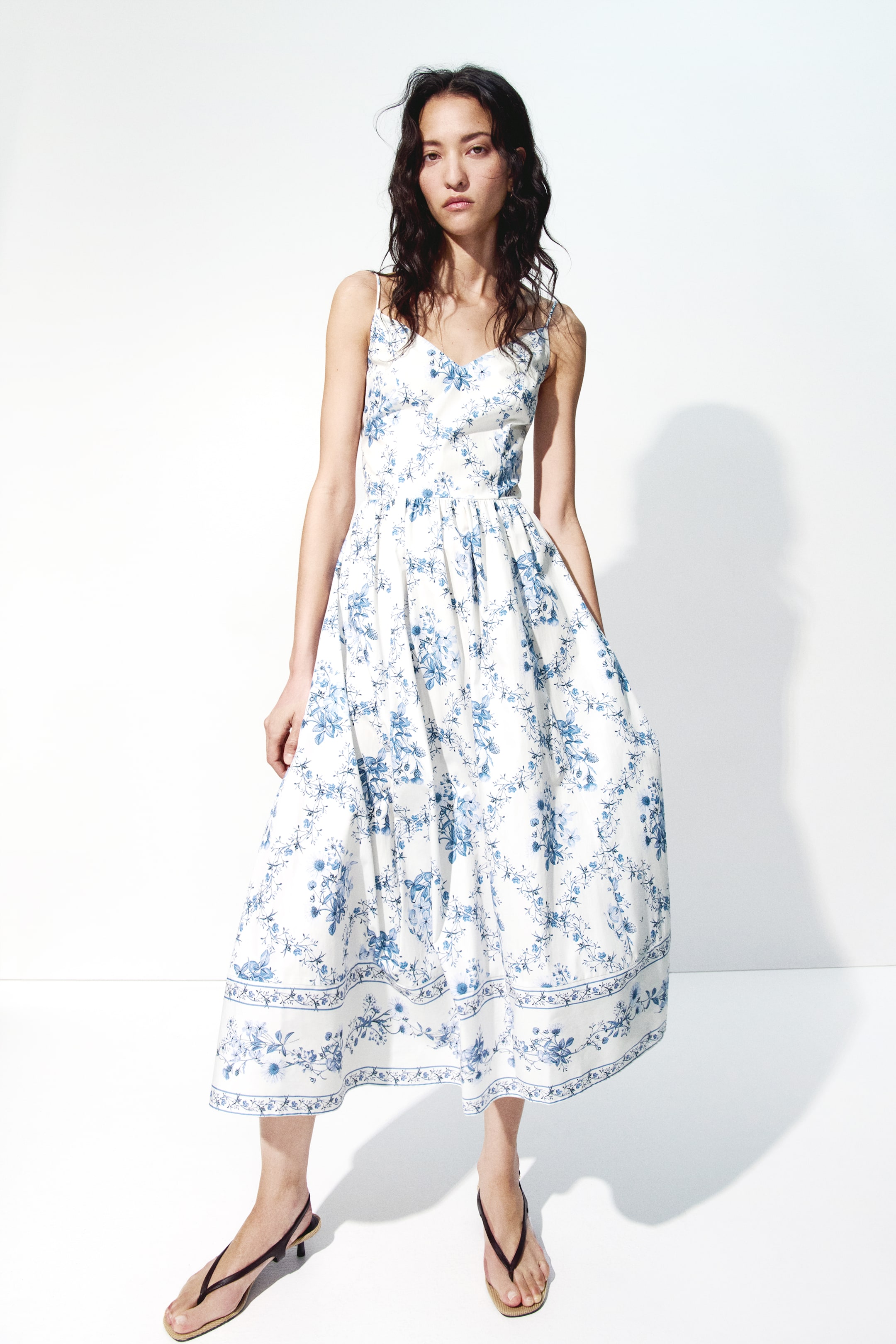 View larger image: A white spaghetti strap midi dress featuring a V-neckline, a fitted bodice, and a gathered waist leading to a full A-line skirt. It displays an all-over blue floral print and a decorative border print at the hem, presenting a front-facing view.