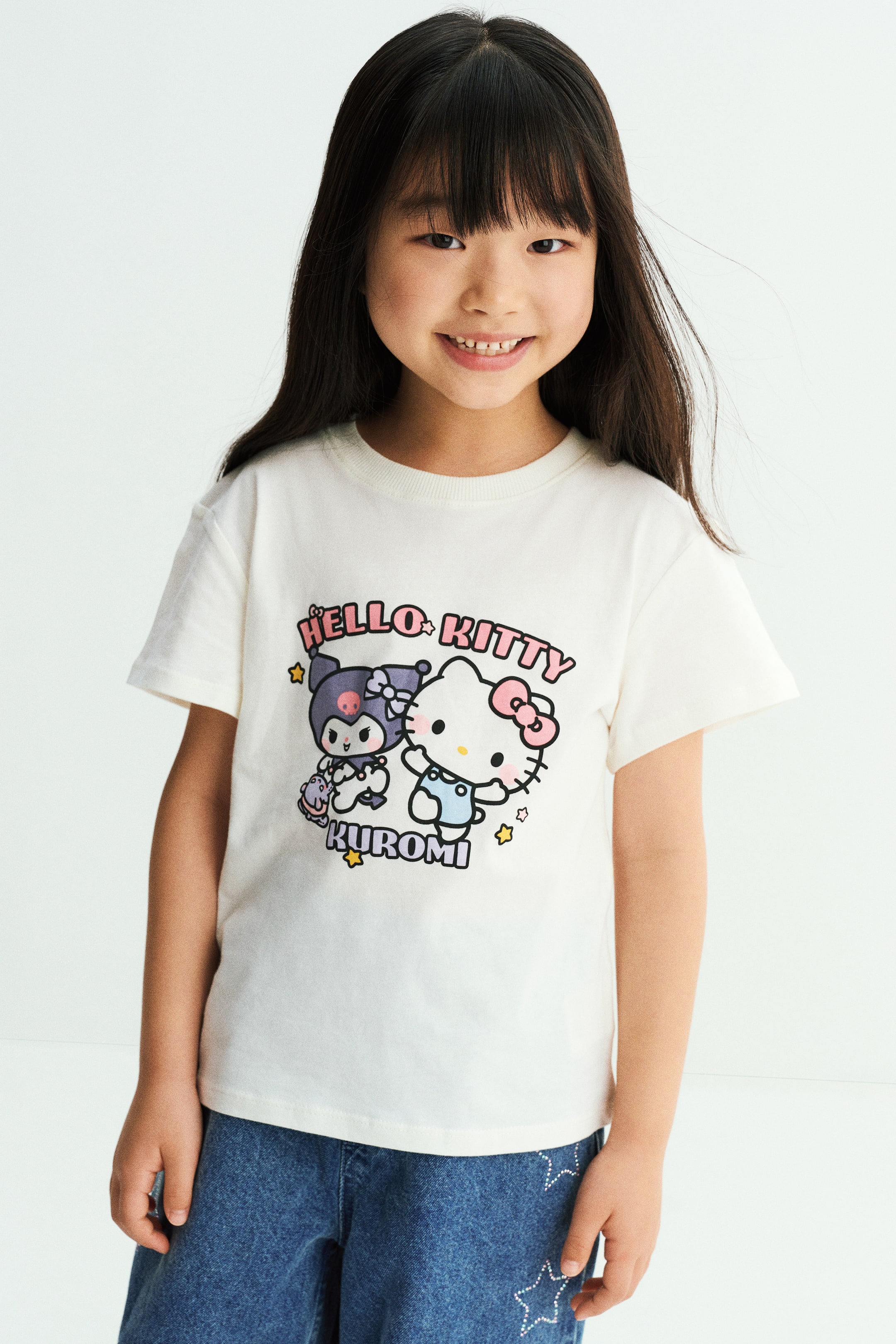 View larger image: A cream-coloured cotton jersey T-shirt featuring a graphic print of Hello Kitty and Kuromi on the front is worn by the model. The T-shirt is designed with a round, rib-trimmed neckline and dropped shoulders, and is paired with blue denim pants.