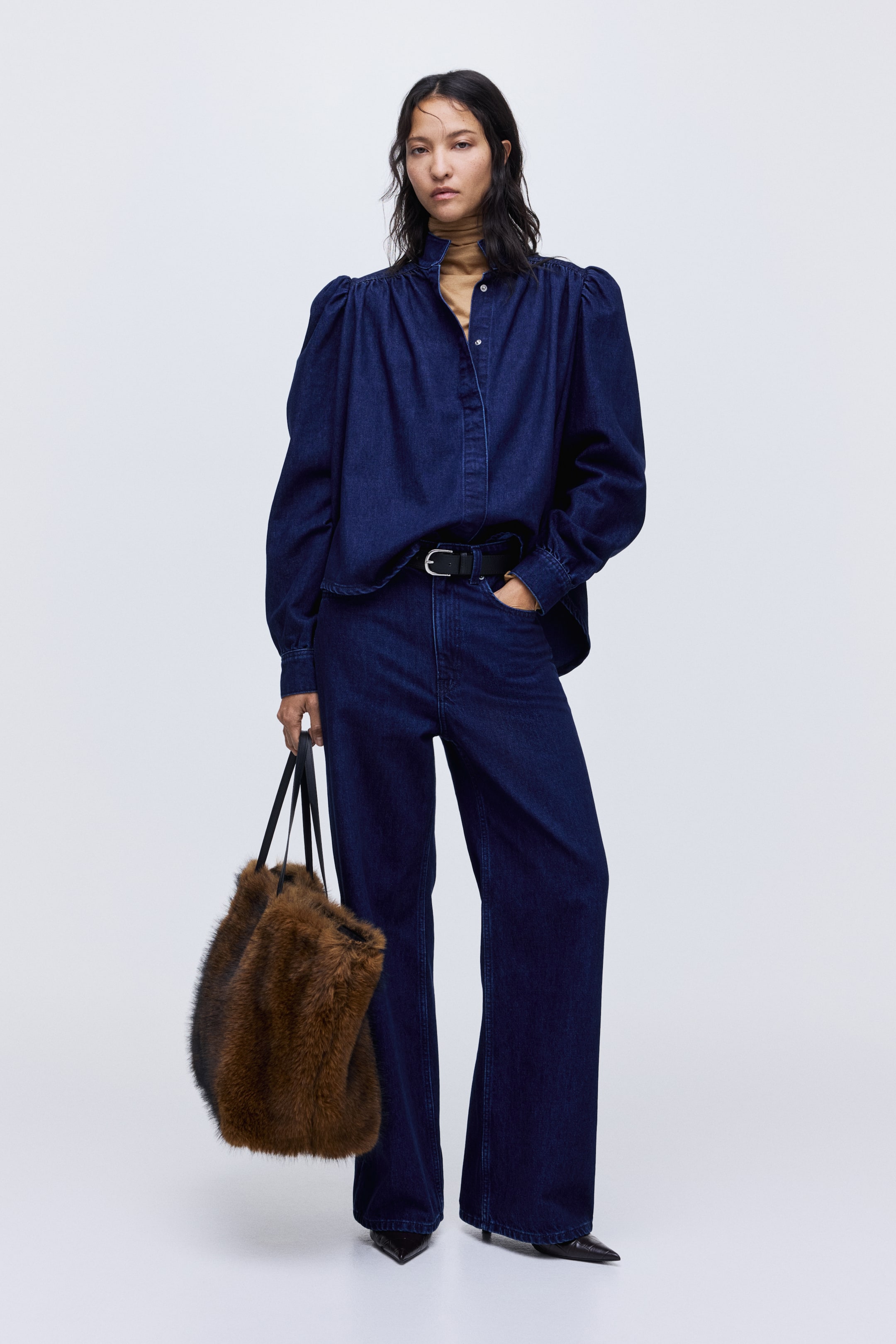 View larger image: A dark blue denim shirt, featuring puffed sleeves and a stand collar, is being presented from a full-front view, layered over a tan turtleneck and paired with matching wide-leg denim pants. A black belt is securing the pants, and a brown faux fur tote bag with black straps is being held.