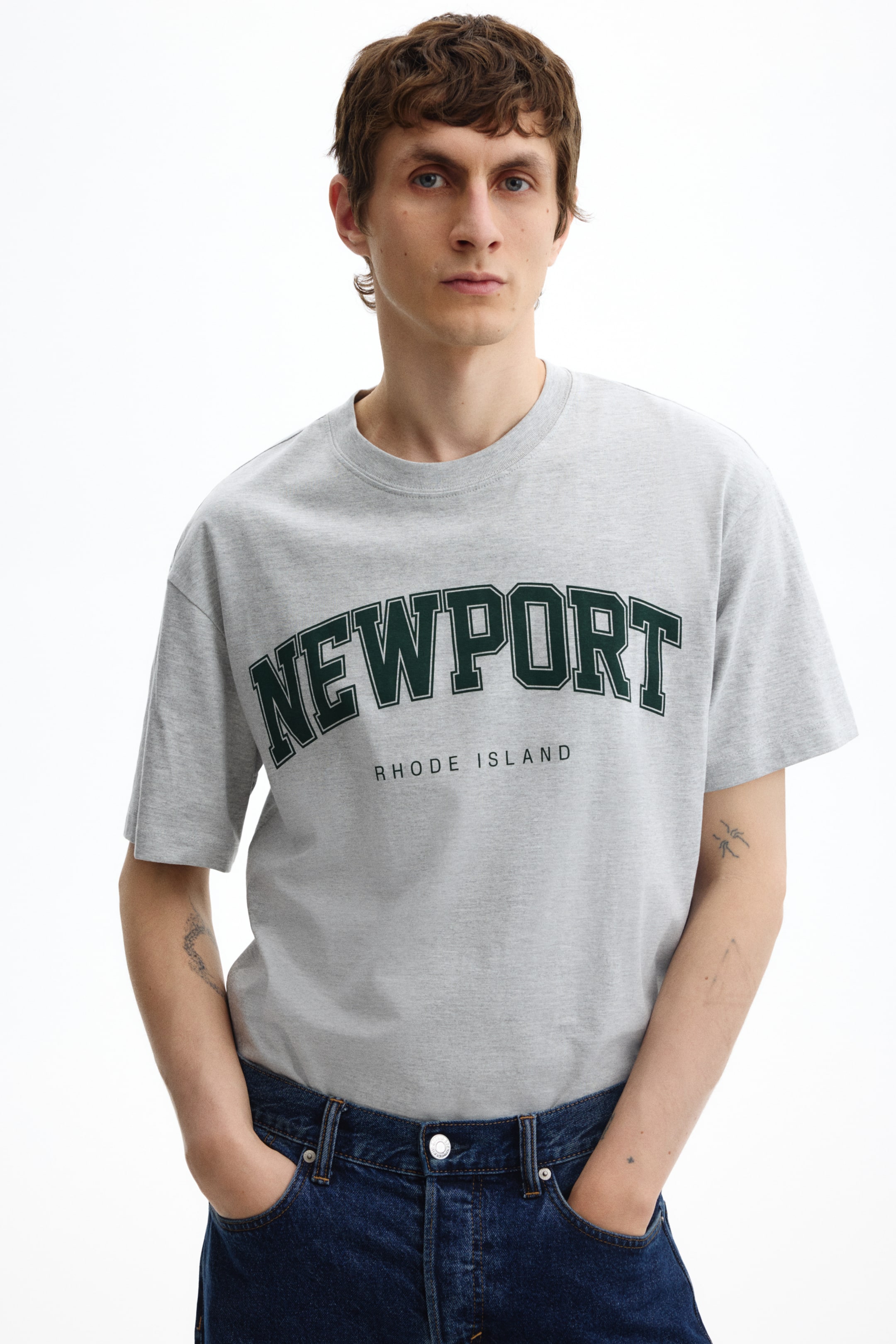 Zobraziť väčší obrázok: Sivé tričko s okrúhlym výstrihom (crew neck) zobrazuje nápis 'NEWPORT' v oblúkových tmavozelených písmenách v kolégiálnom štýle, pod ktorým je vytlačený nápis 'RHODE ISLAND'. Má voľný strih, krátke rukávy a je zobrazené zepredu.