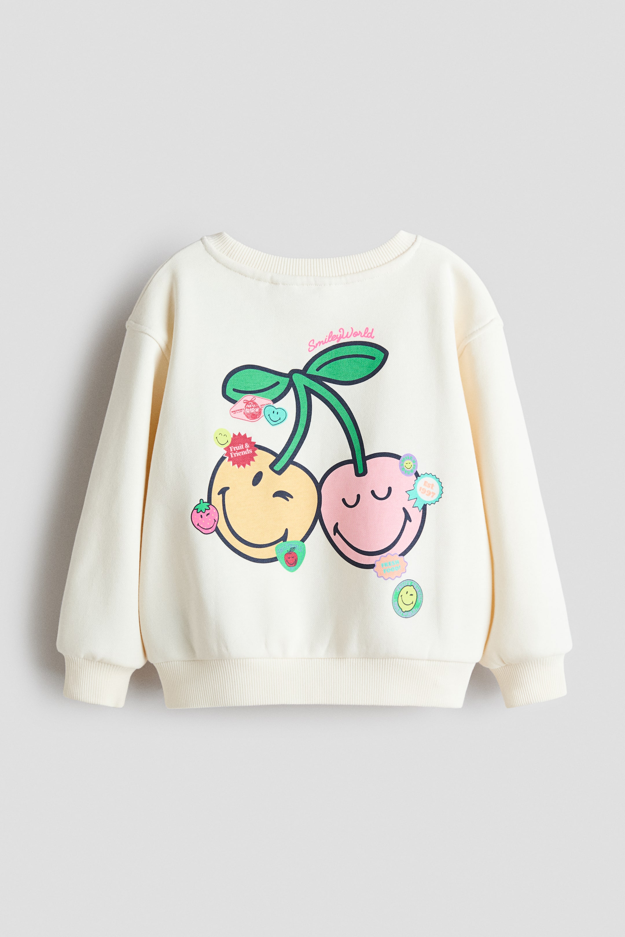 View larger image: A cream sweatshirt is presenting its FRONT, featuring a large cherry motif with yellow and pink smiley faces as the cherries, surrounded by smaller fruit and smiley face stickers. It is designed with dropped shoulders, long sleeves, and ribbed detailing at the crew neckline, cuffs, and hem.