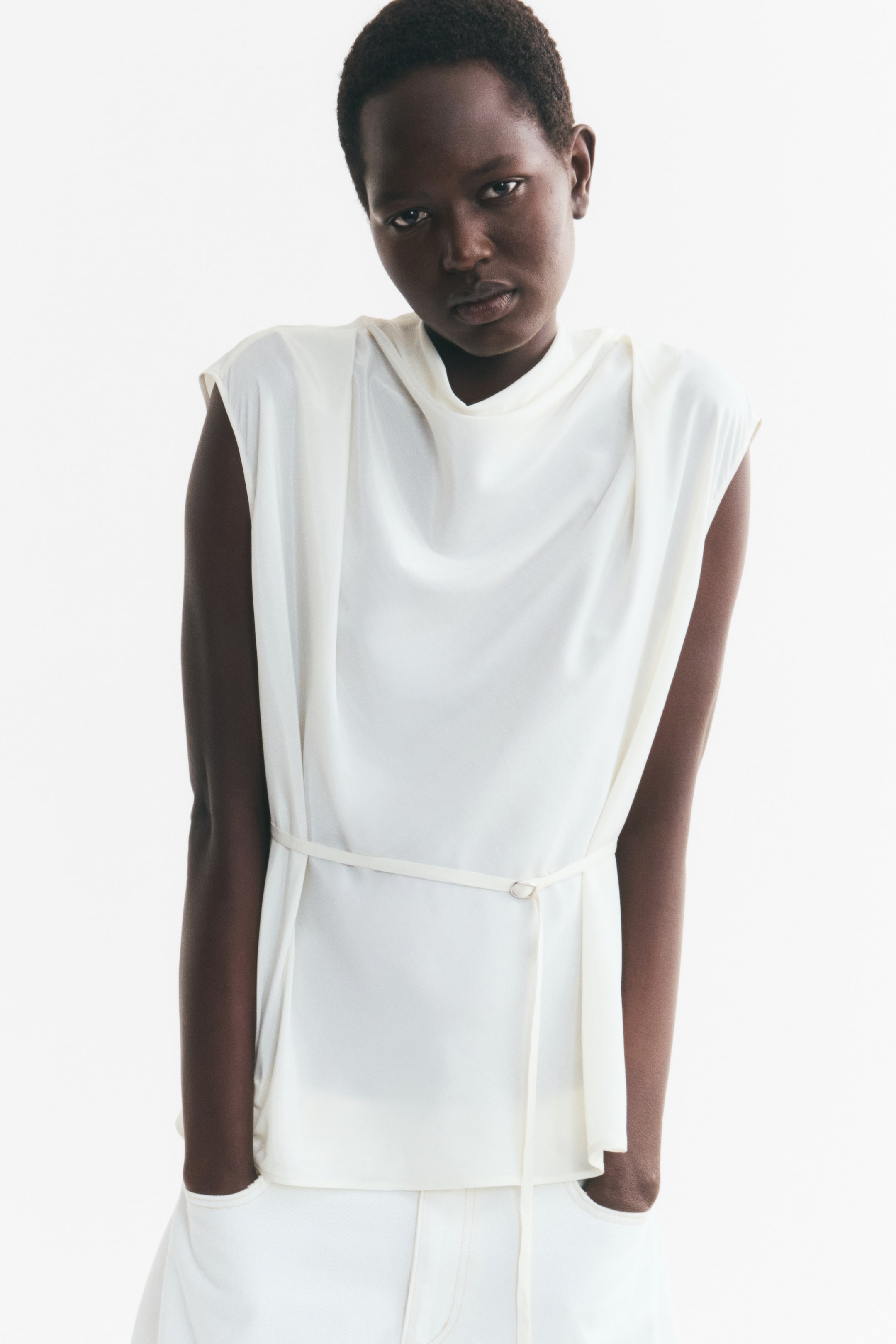 View larger image: The model is wearing an off-white sleeveless top with a draped cowl neckline and a slim matching belt. The top features structured, extended shoulders and is paired with matching white trousers.