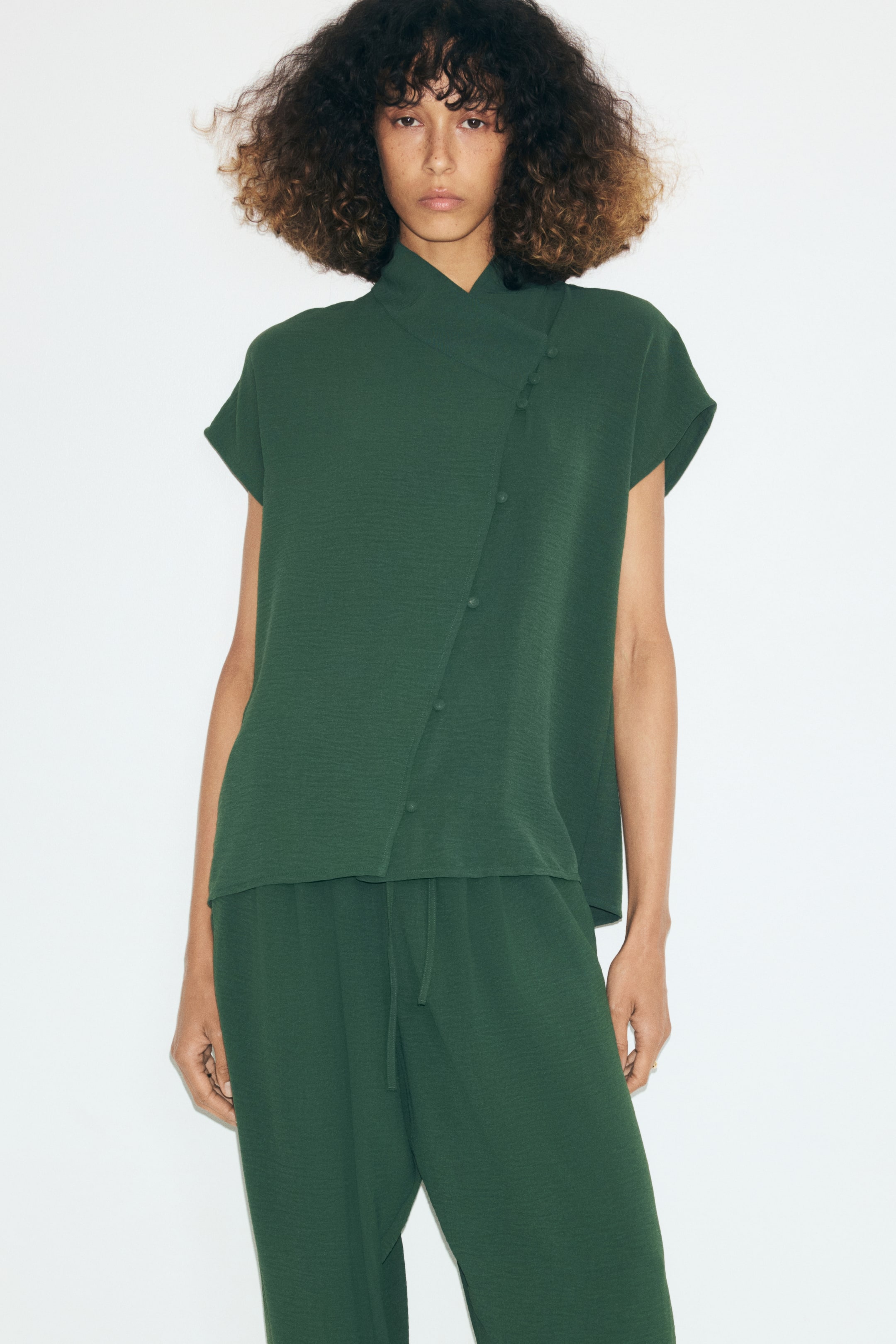 Ampliar la imagen: La modelo lleva una blusa tejida en verde oscuro con escote cruzado alto, mangas tipo cap y una abertura frontal descentrada con botones forrados. La blusa se combina con un pantalón a juego en verde oscuro.
