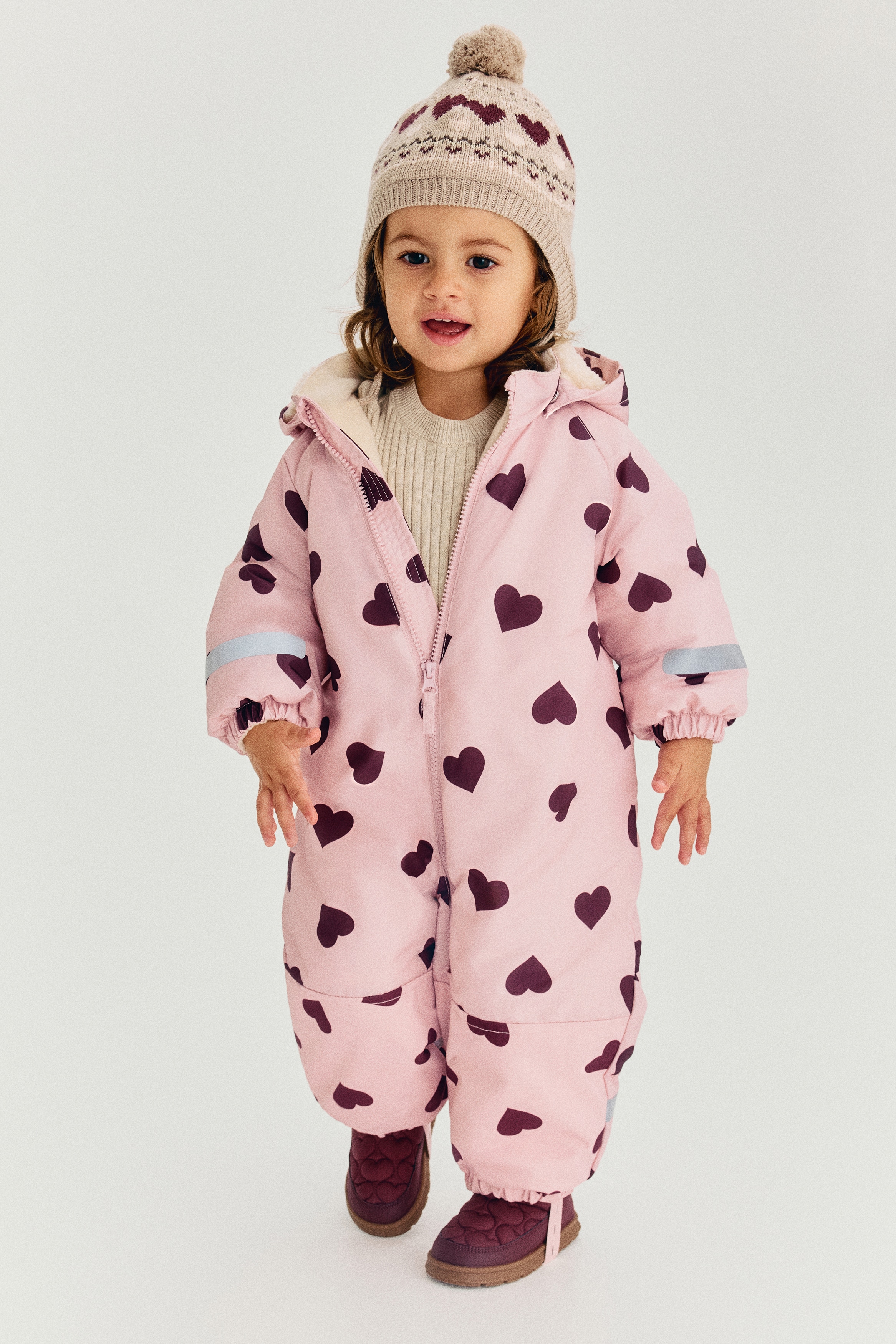 A padded baby all-in-one suit is showcasing a light pink base adorned with a dark red heart pattern, viewed from the front. It features a full-length front zipper, a detachable hood and stand-up collar with a cream fleece lining, elasticated cuffs with a reflective strip on the left arm, and elasticated hems with detachable foot straps.