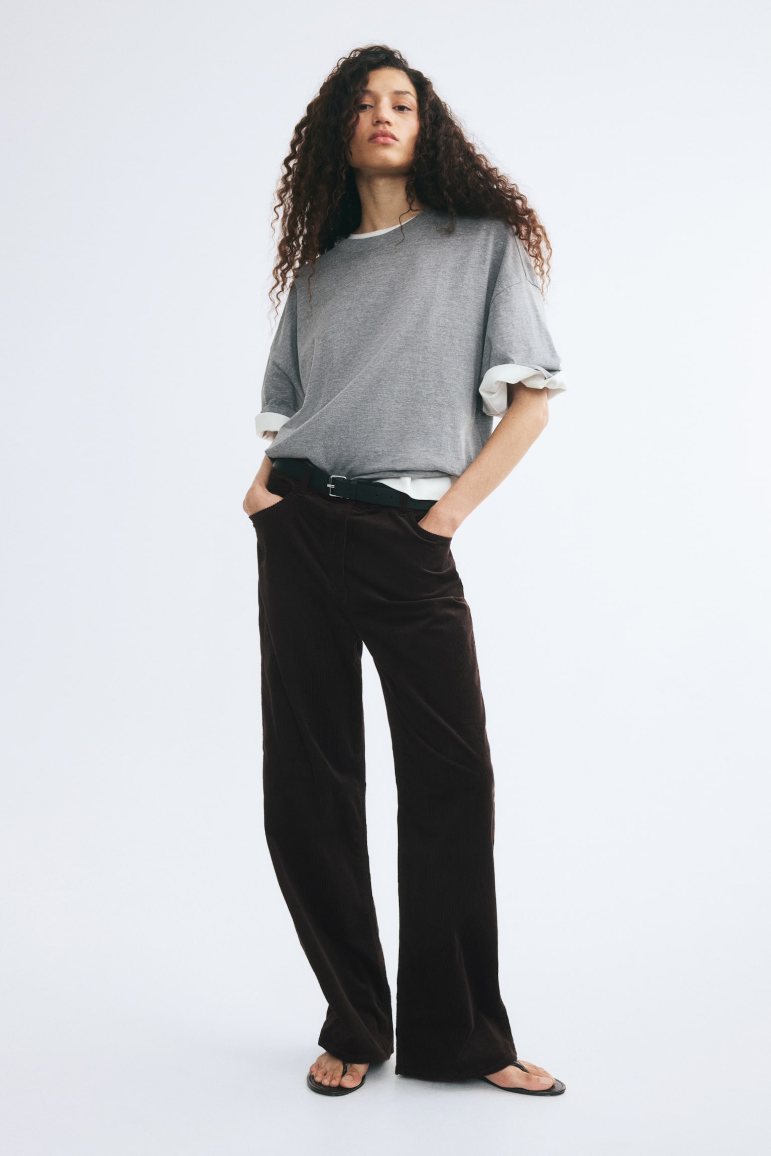 Dark brown corduroy trousers featuring straight legs and a high waist are being worn by the model, who is standing with hands resting in the front pockets. The trousers are secured with a black buckled belt and styled with a grey t-shirt tucked into the waistband. - 1
