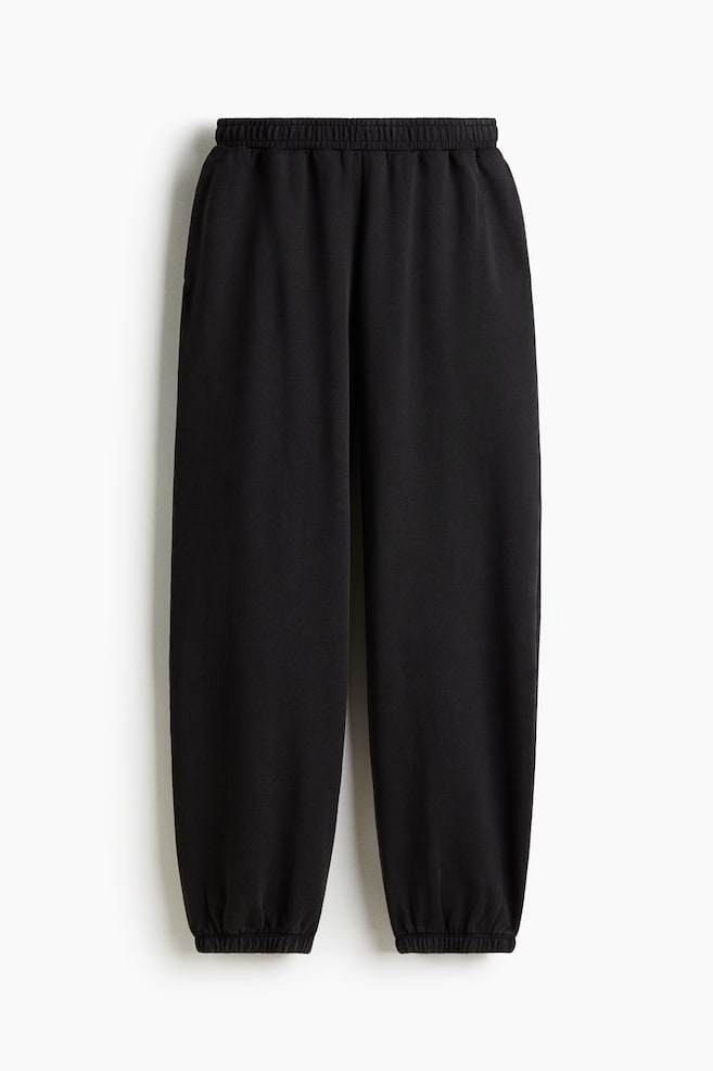 Black sweatpants are being presented from the front, featuring an elasticated waistband, subtle side seam pockets, and gathered elastic cuffs at the ankles. - 1