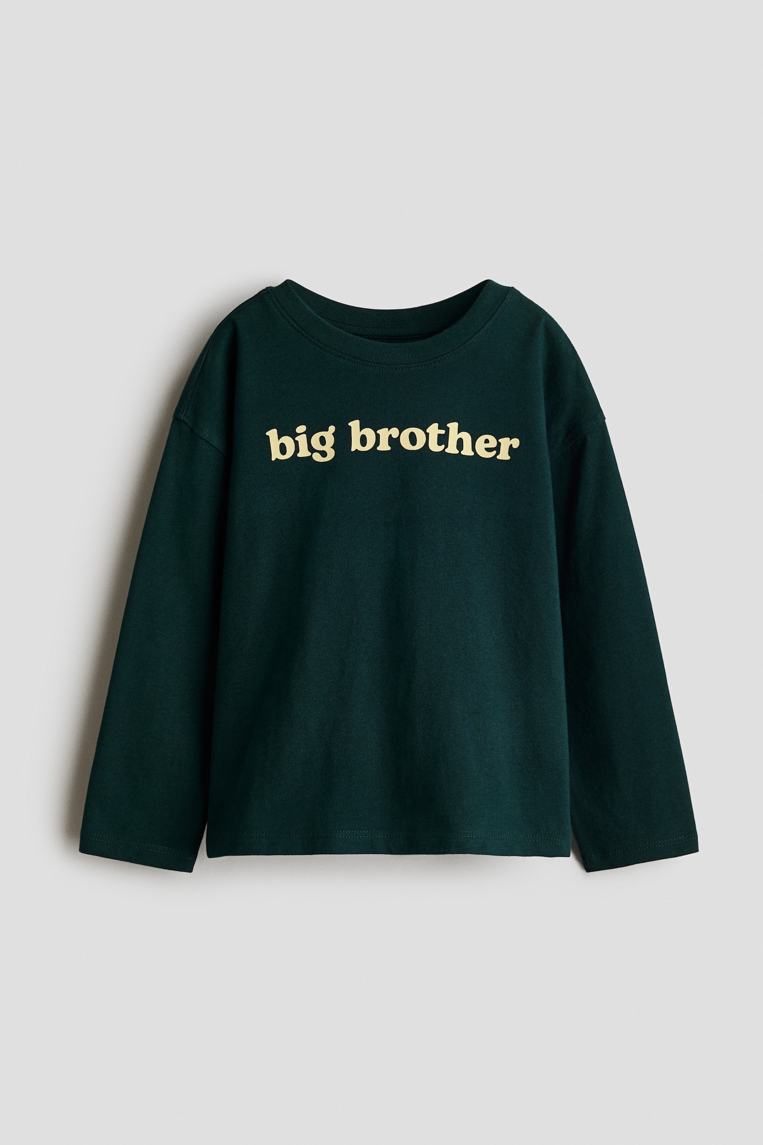 A front view of a dark green, long-sleeve cotton jersey T-shirt displaying the text "big brother" printed in a pale yellow lowercase font across the chest. The T-shirt features dropped shoulders and a round crew neckline with a ribbed trim. - 1