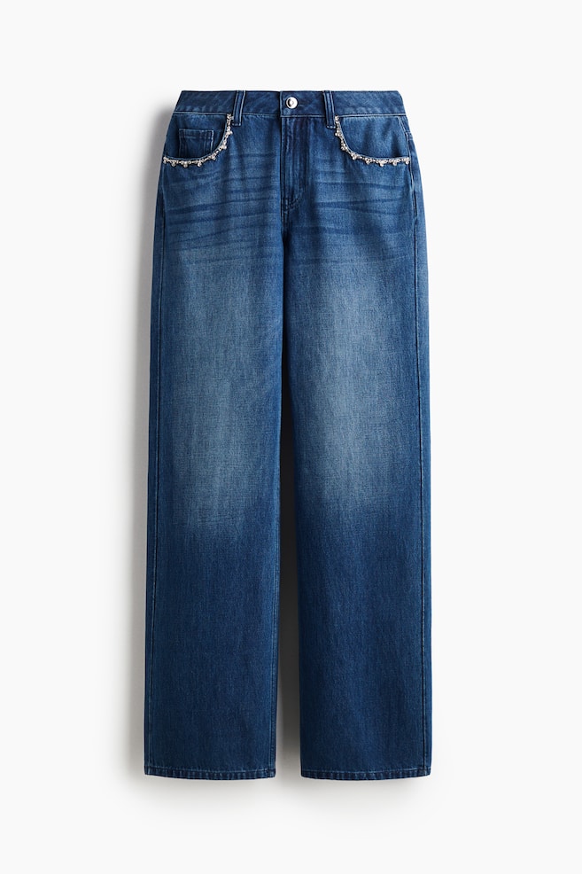 Mavi denim kot pantolonlar önden sunuluyor, uyluklarda açık yıkama ve düz paça kesimi ile dikkat çekiyor. Bel kısmını tek bir gümüş tonlu düğme sabitliyor ve ön ceplerin üst kenarları boyunca gümüş tonlu boncuklu bir zincirle süsleniyor. - 1