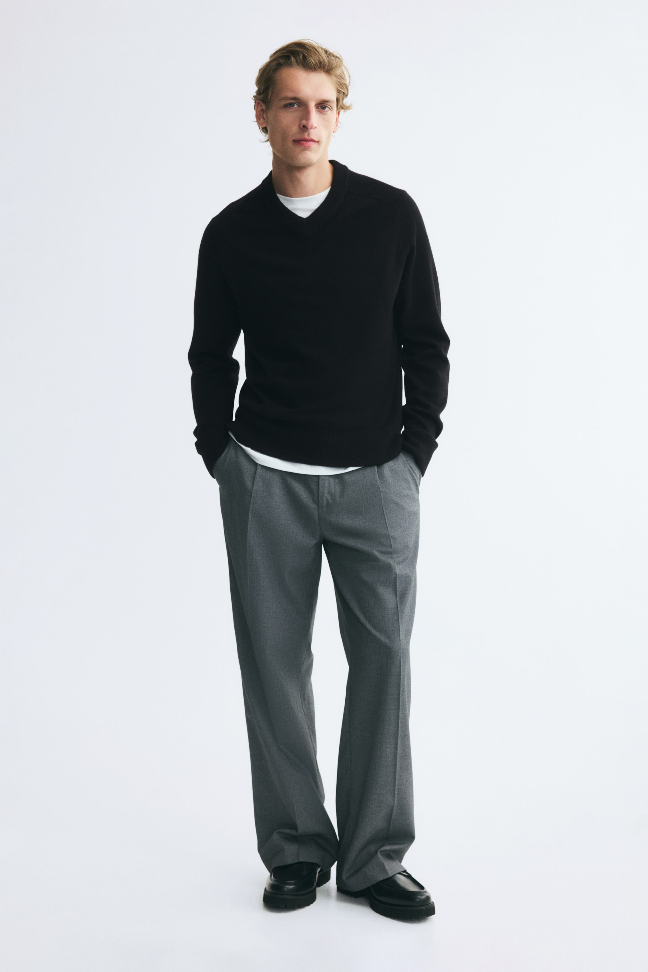 View larger image: A black knit v-neck sweater is being worn by the model, layered over a white t-shirt and paired with relaxed-fit grey trousers. The long-sleeved sweater features ribbed detailing along the neckline, cuffs, and hem.