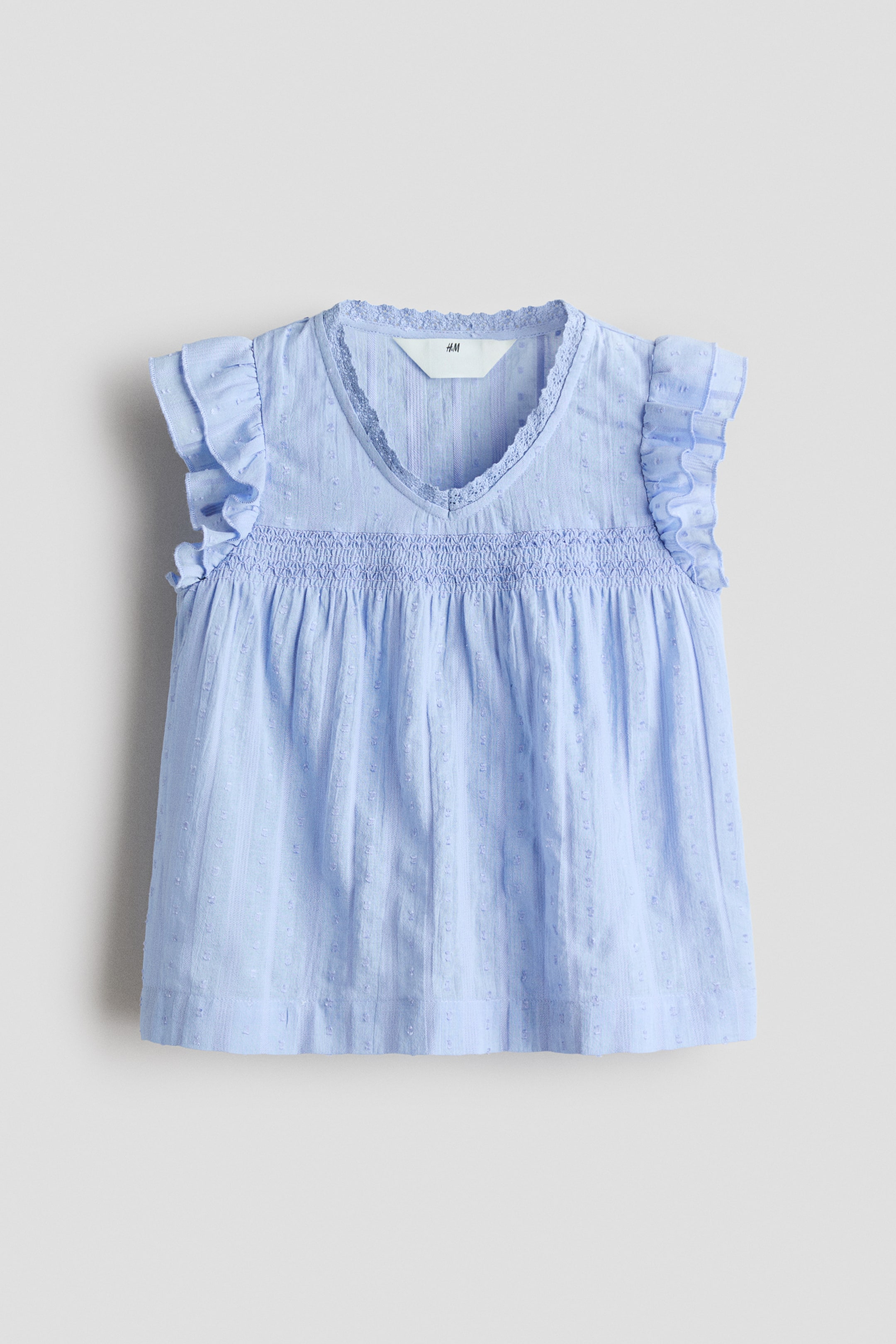 View larger image: A light blue blouse is showcasing its front view, highlighting an airy weave with small textured dots. It is crafted with a lace-trimmed V-shaped neckline, double-layered flutter sleeves, smocking across the front, and a gently flaring hem.