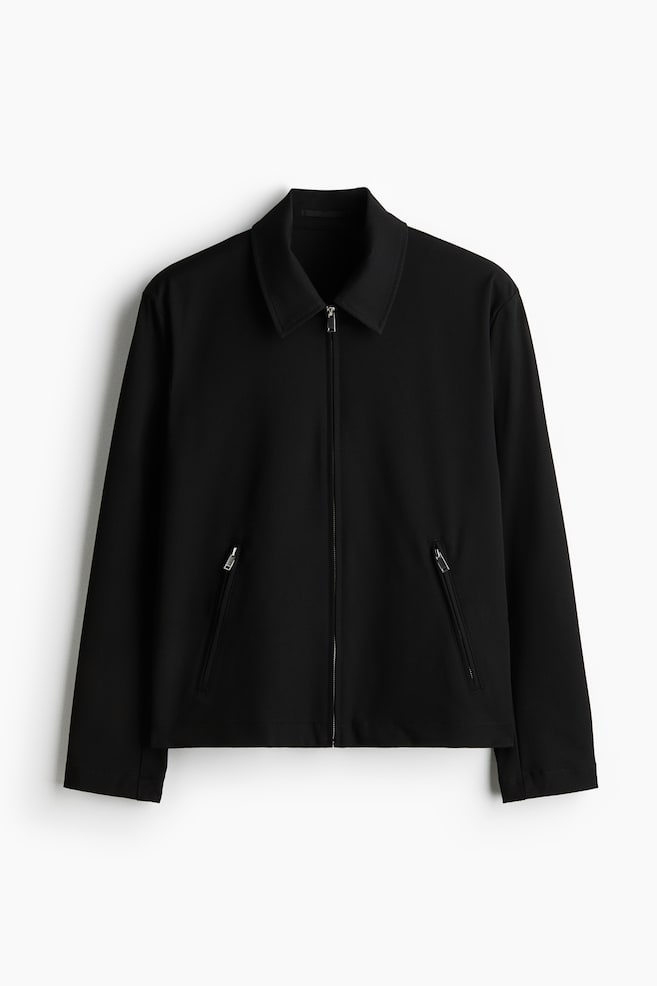 A black jacket is displayed from the front, featuring a pointed collar and a full-length central zipper. It is designed with two angled zipper pockets on the lower front and long sleeves ending in straight cuffs. - 1