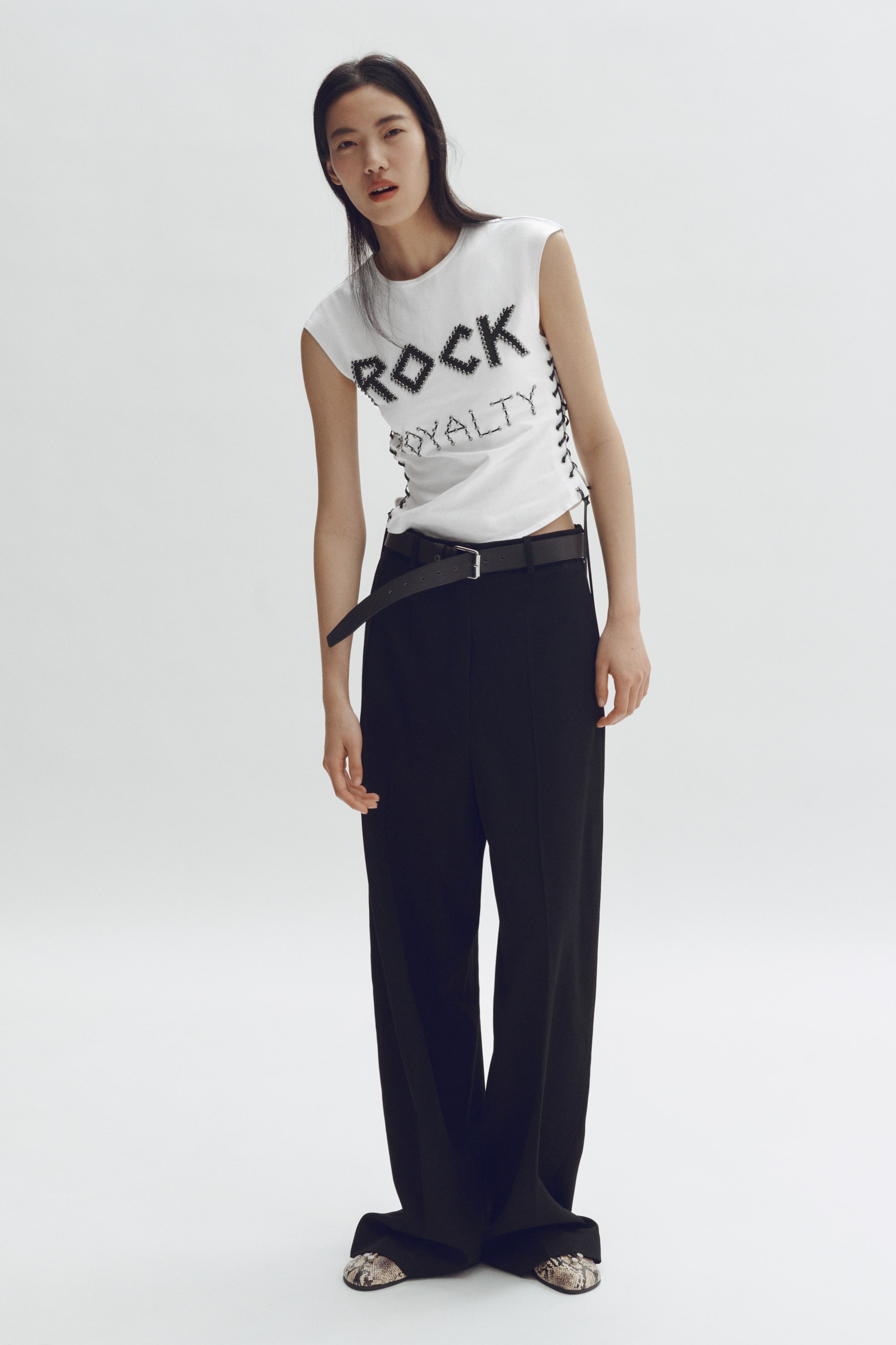 View larger image: A white sleeveless top, adorned with black "ROCK ROYALTY" text and side lacing details, is teamed with black wide-leg trousers, a black belt with a silver buckle, and pointed-toe shoes featuring a snakeskin pattern.