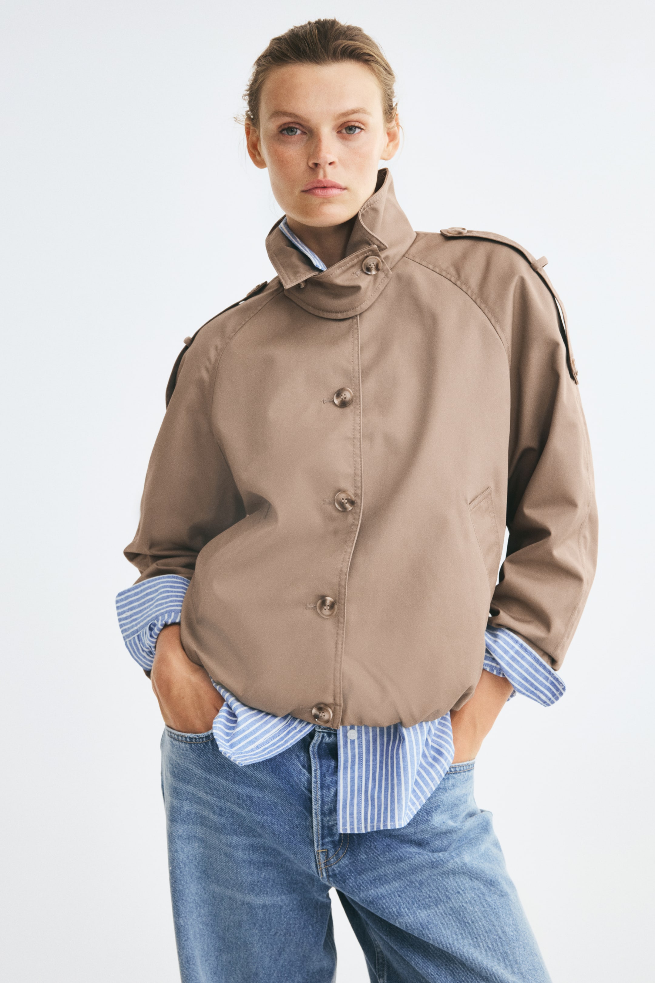 Xem ảnh lớn hơn: Người mẫu đang mặc một chiếc áo khoác blouson cỡ rộng màu nâu chất liệu twill, nổi bật với cổ cao kiểu funnel có quai cài cổ bằng nút, tab vai cài nút và gấu bo chun tạo dáng phồng. Áo khoác được cài nút phía trước và phối lớp bên ngoài với áo sơ mi sọc xanh trắng cùng quần jeans xanh.