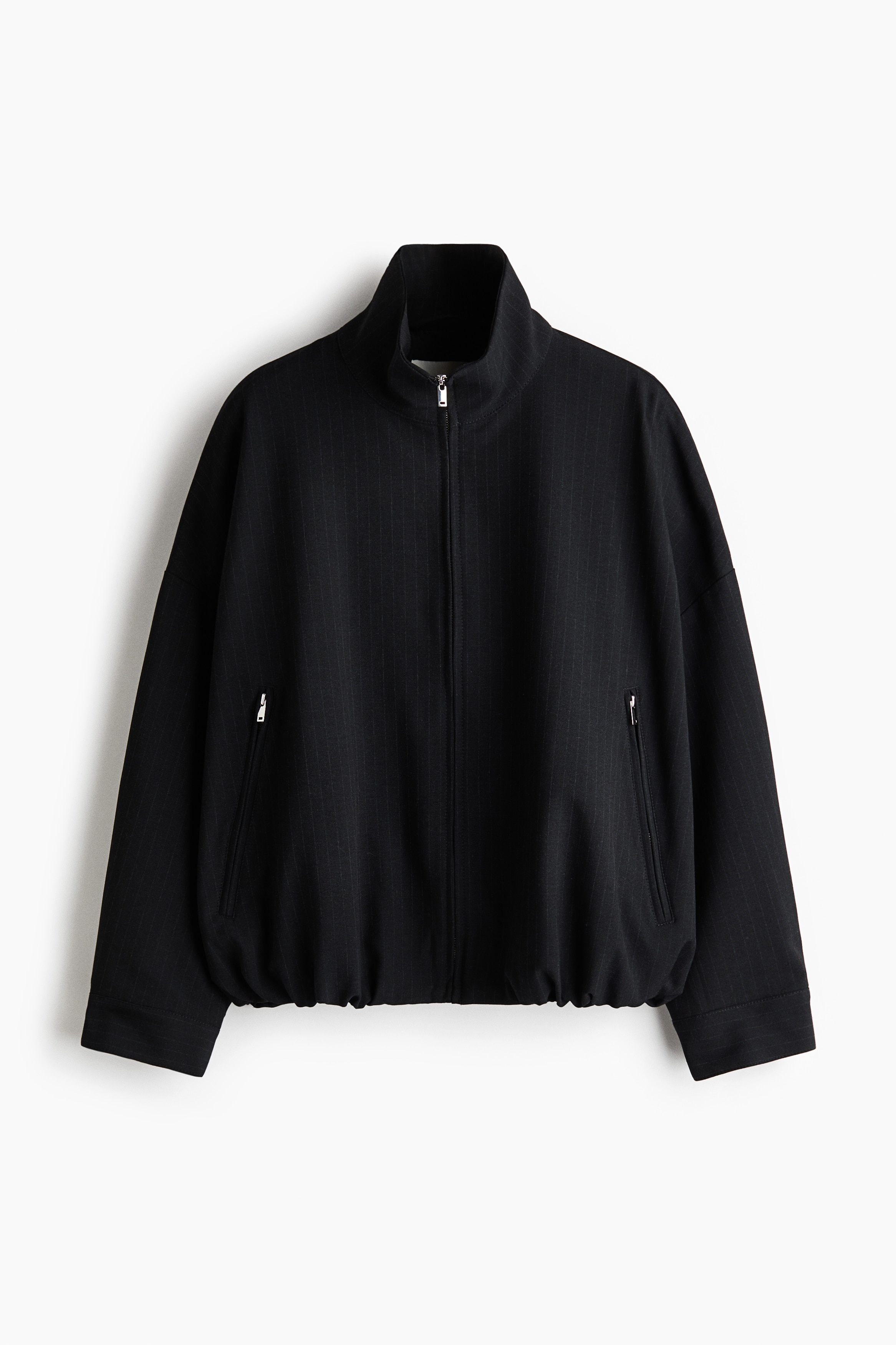 The model is wearing an oversized black jacket with a stand collar, front zip fastening, and an elasticated bubble hem. The jacket is styled with a white T-shirt, matching black trousers, and a black shoulder bag.