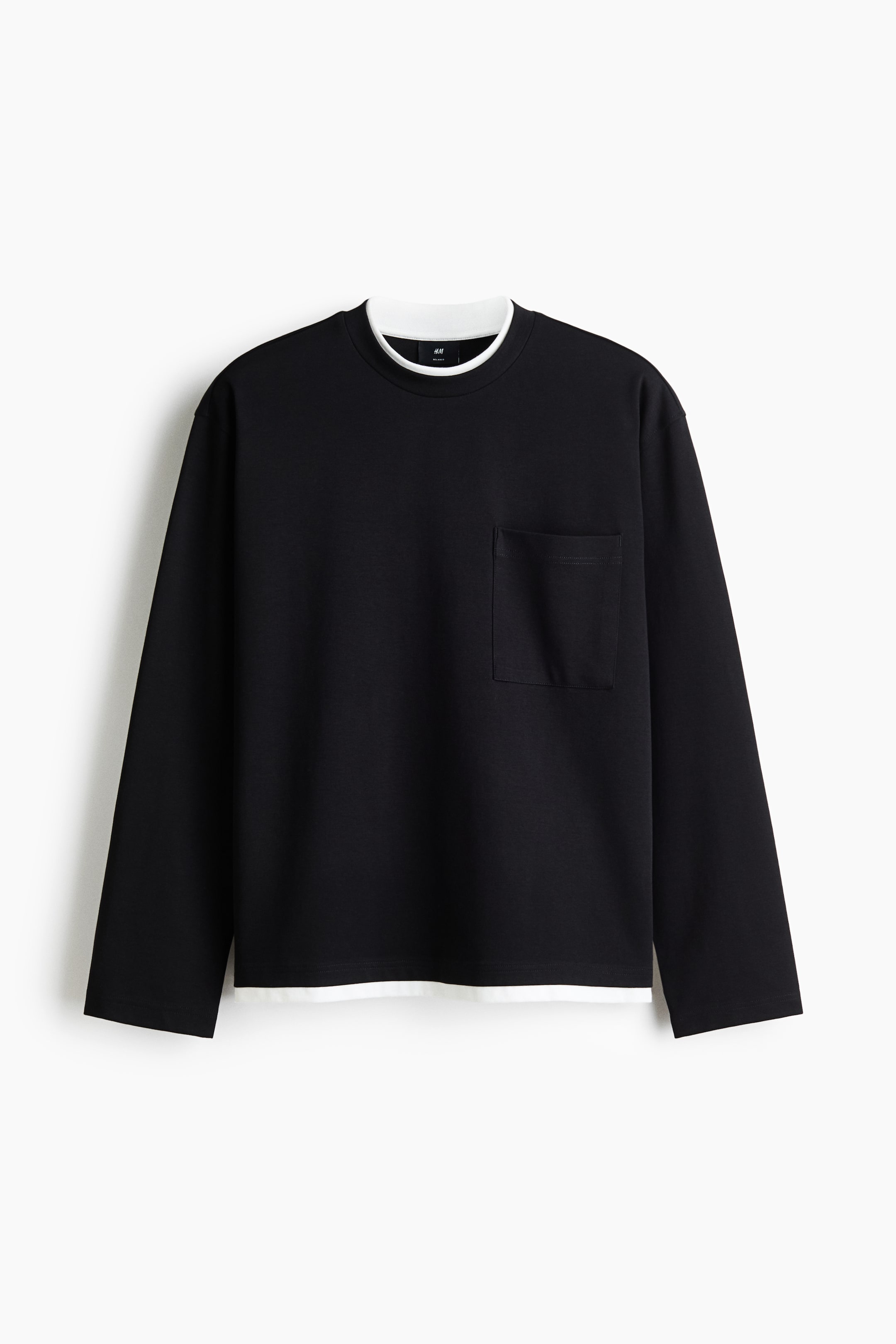 Größeres Bild ansehen: Ein schwarzes Langarmshirt ist von vorne zu sehen, mit einem runden Halsausschnitt und einer rechteckigen Brusttasche auf der linken Seite. Eine weiße Schicht ist unter dem Halsausschnitt und am unteren Saum sichtbar.