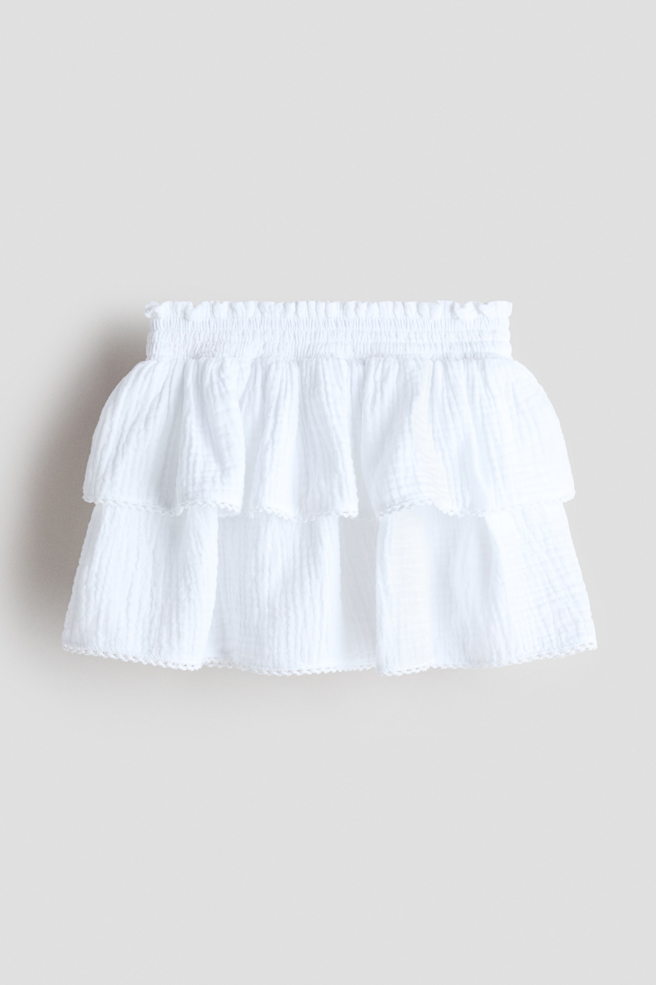 View larger image: A white front-facing skirt featuring a shirred elastic waistband and two gathered tiers of crinkled fabric. Each tier is finished with a delicate picot trim along its hem.